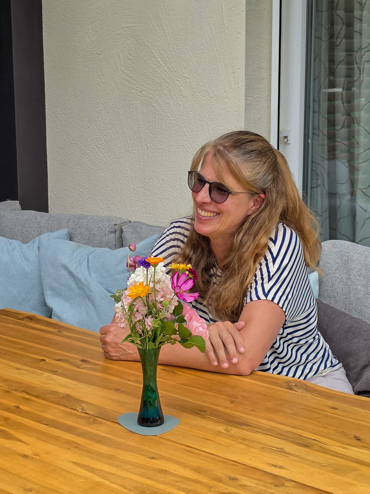 A smiling woman with sunglasses sits at a wooden table with a vase of colorful flowers on it.