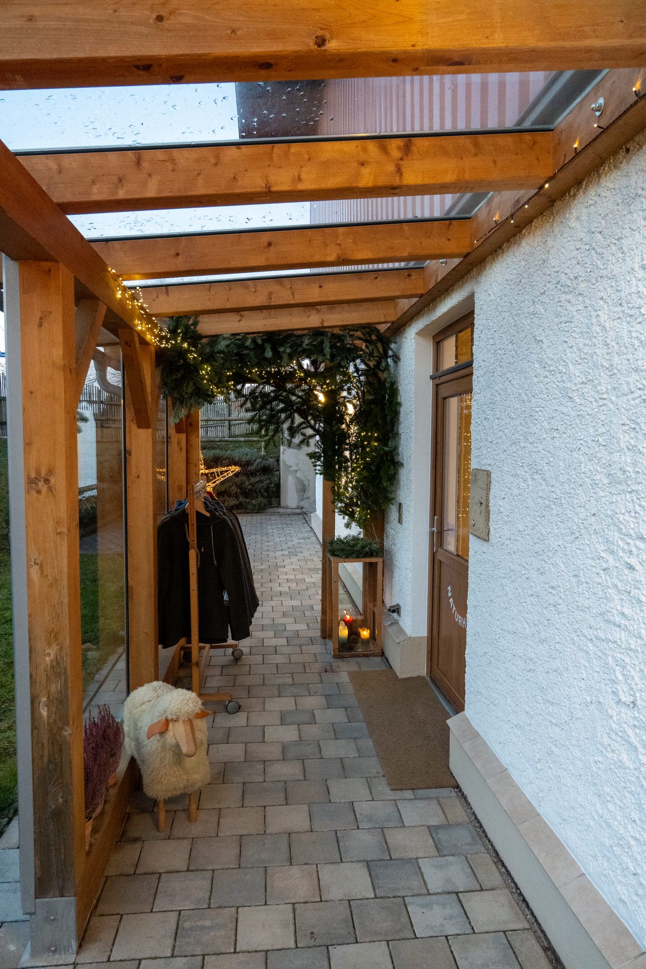 Festive wooden-covered walkway with fairy lights, evergreen decor, a sheep figure, and a door.