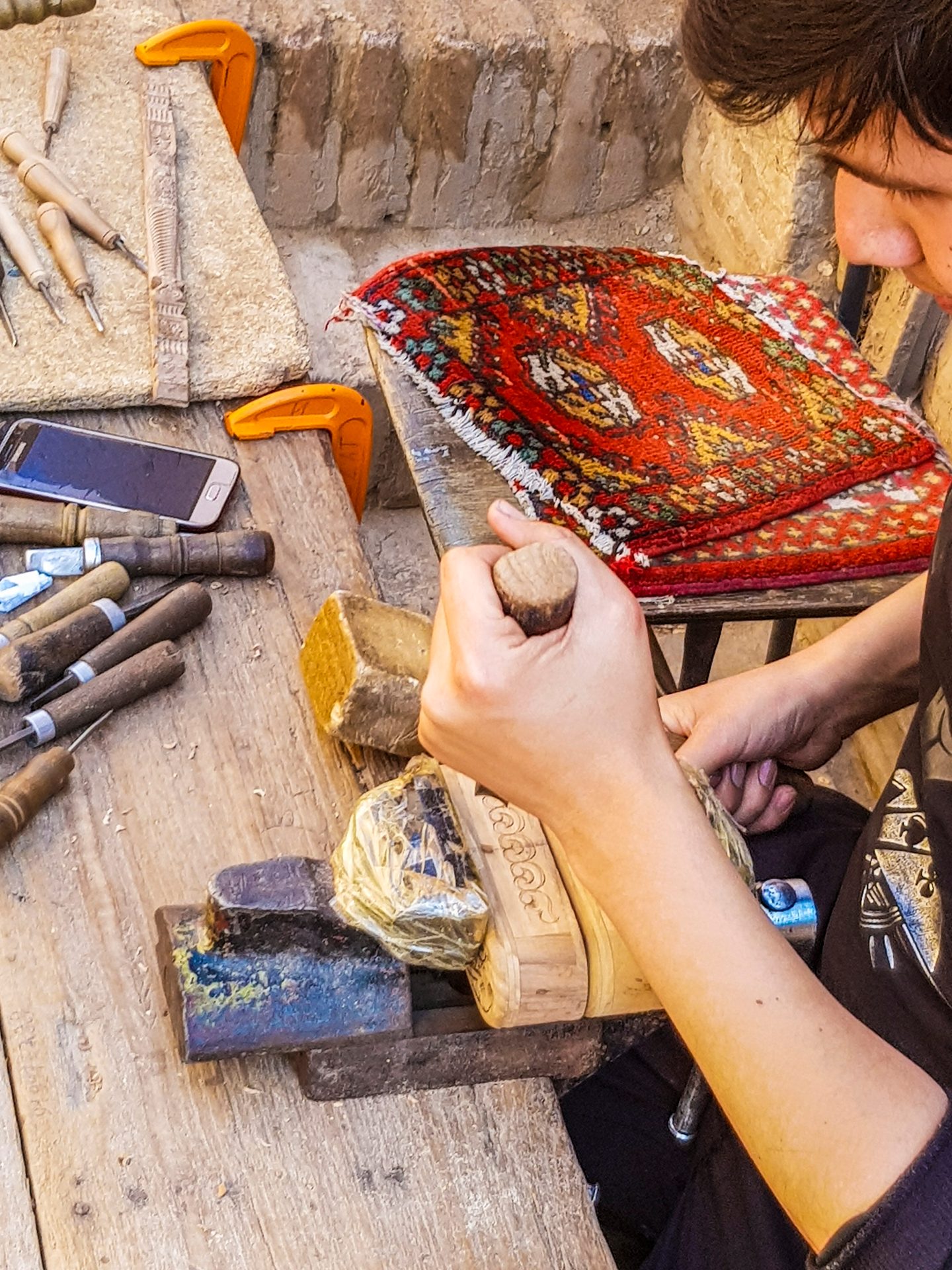 Hands carving intricate patterns into wood with tools on a workbench, next to a smartphone and rug.