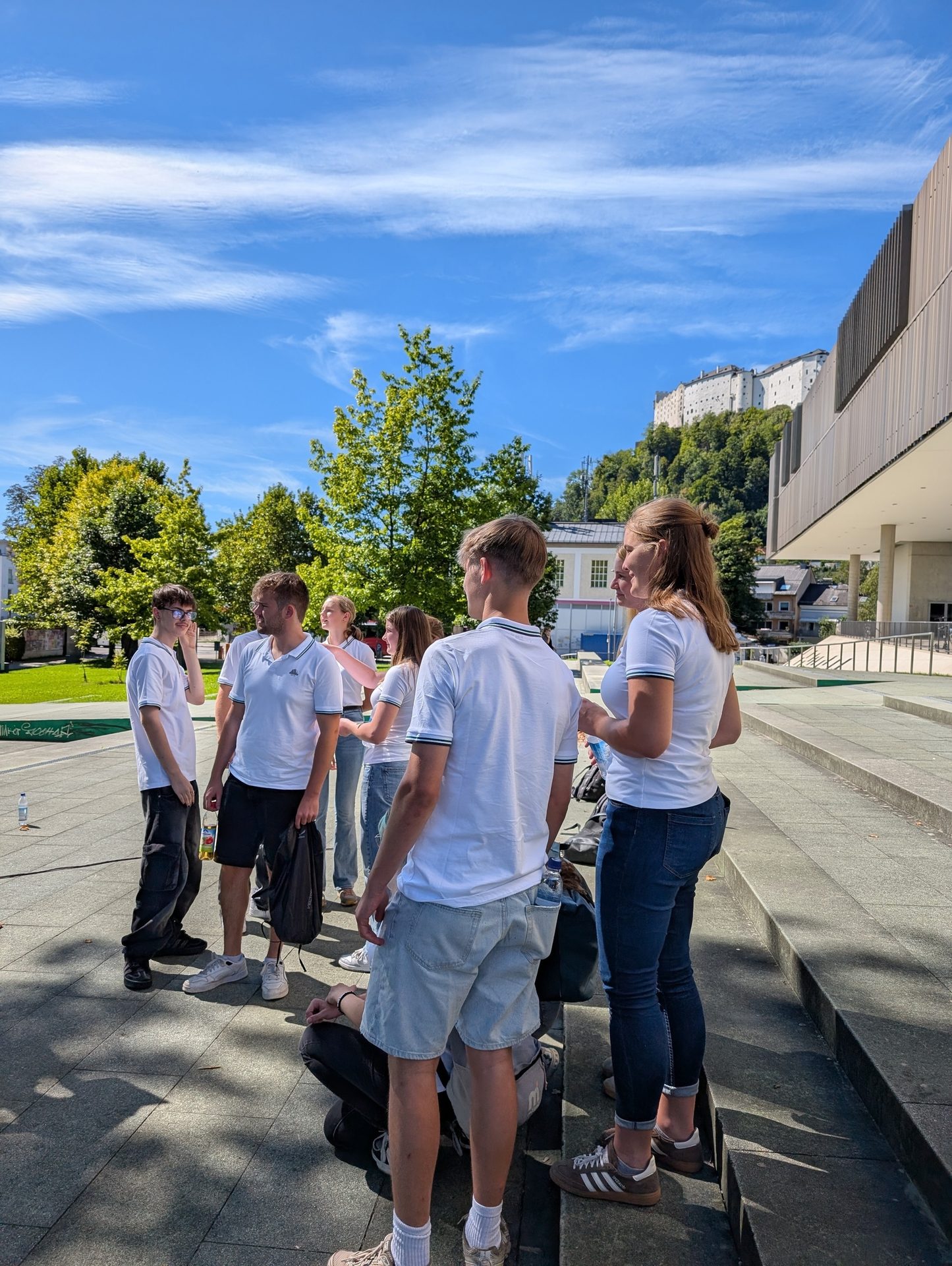 A group of young people gathered outdoors on a sunny day, with a castle and modern building in the background.