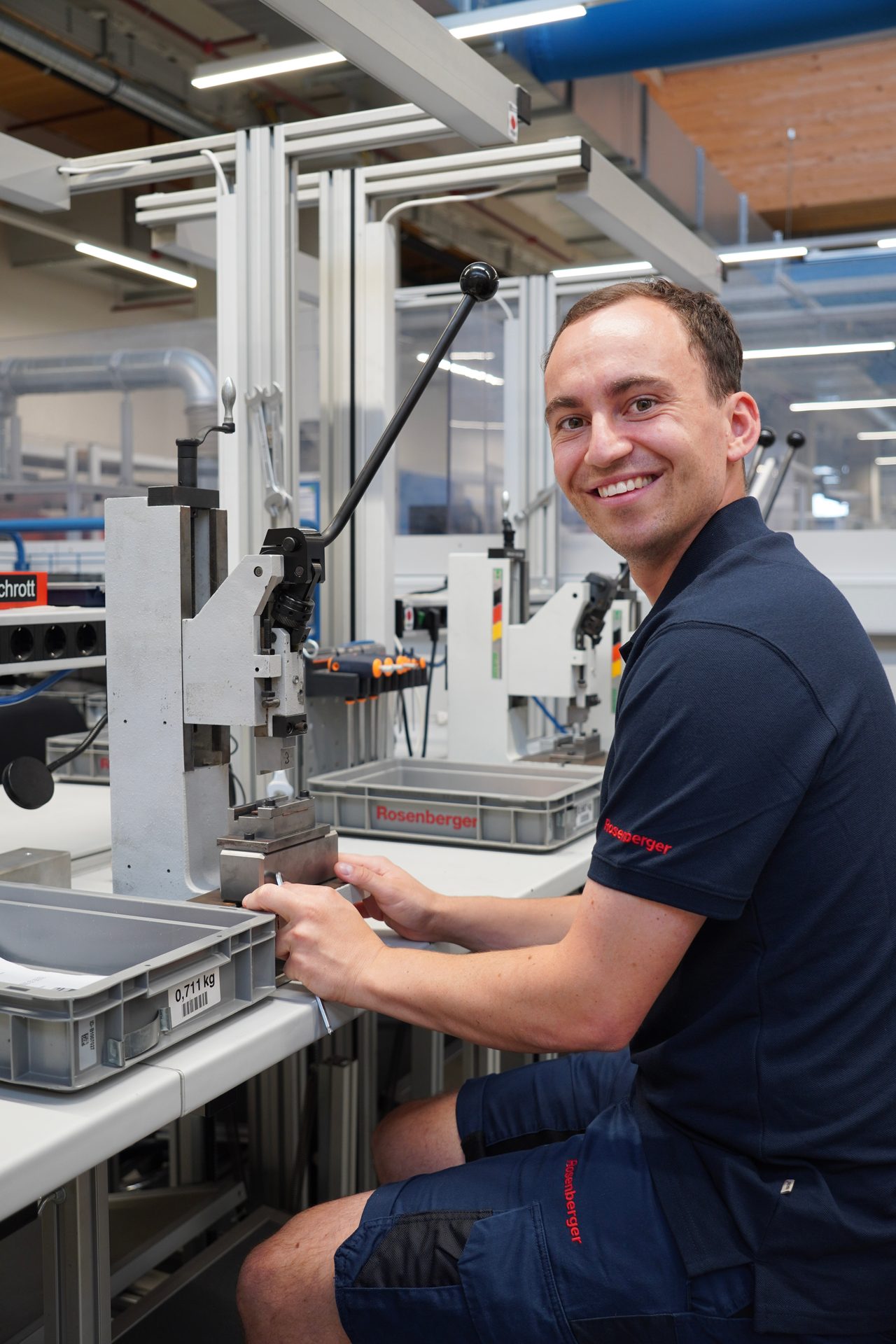 A smiling man in a dark blue polo shirt operates a press machine in a factory, looking at the camera.