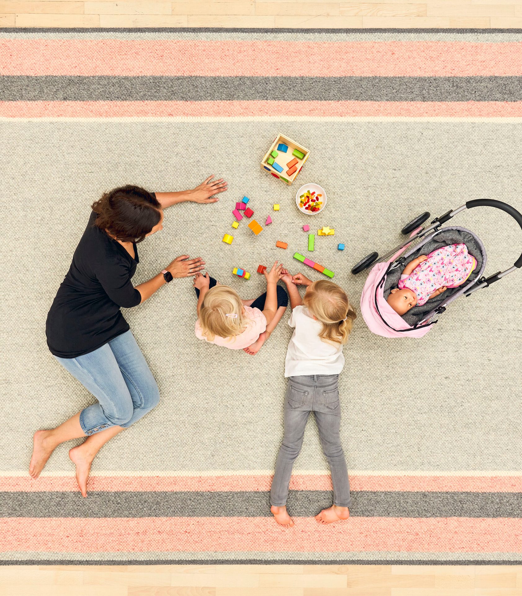 Overhead view of a woman and two girls playing with toys and a doll stroller on a striped rug.