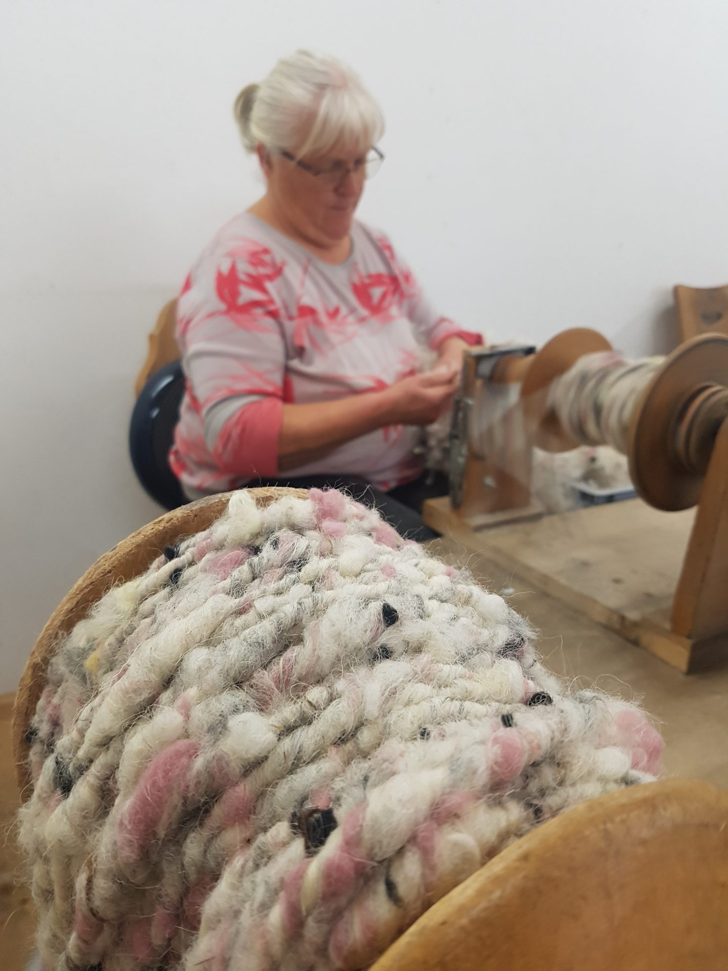 Close-up of pink and white textured yarn on a spinning wheel, with a woman spinning in the background.