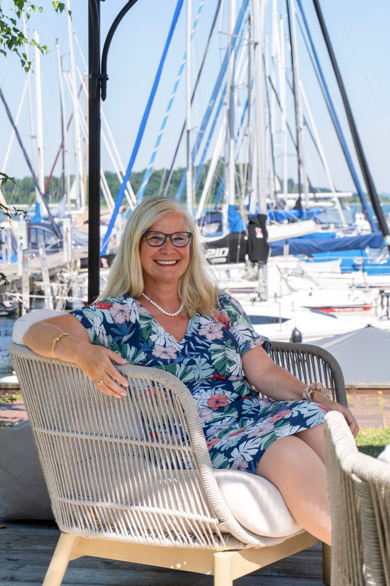 Smiling woman with glasses sits in a chair by a marina with boats.