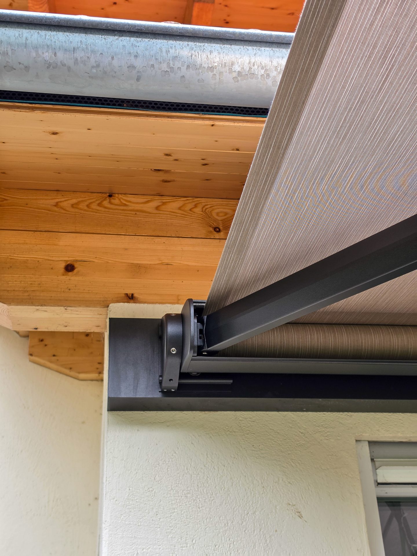An awning mechanism with beige fabric extended, mounted on a wall below a metal gutter and wooden beams.
