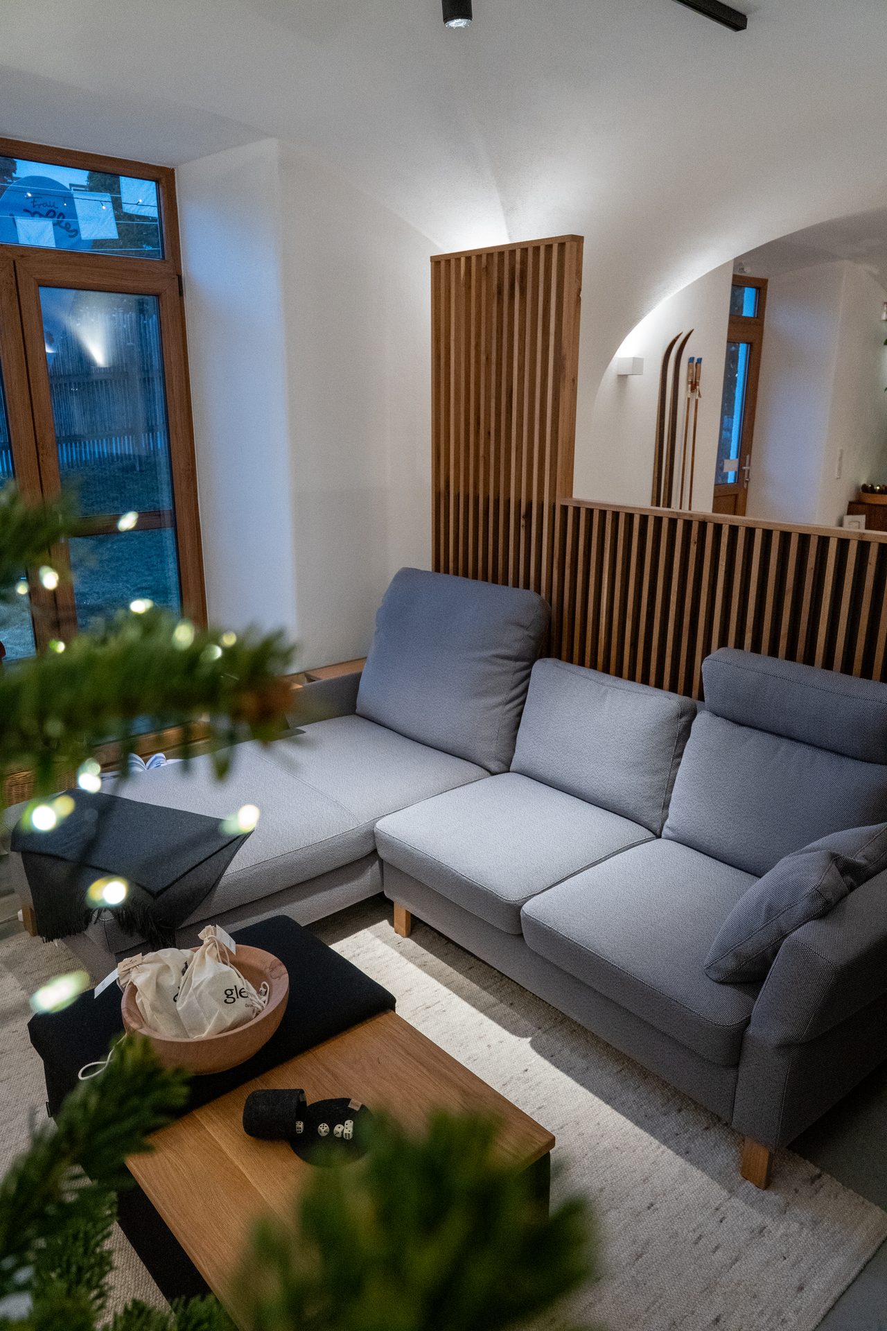 Cozy modern living room with grey sectional sofa, wooden coffee table, and Christmas tree lights.