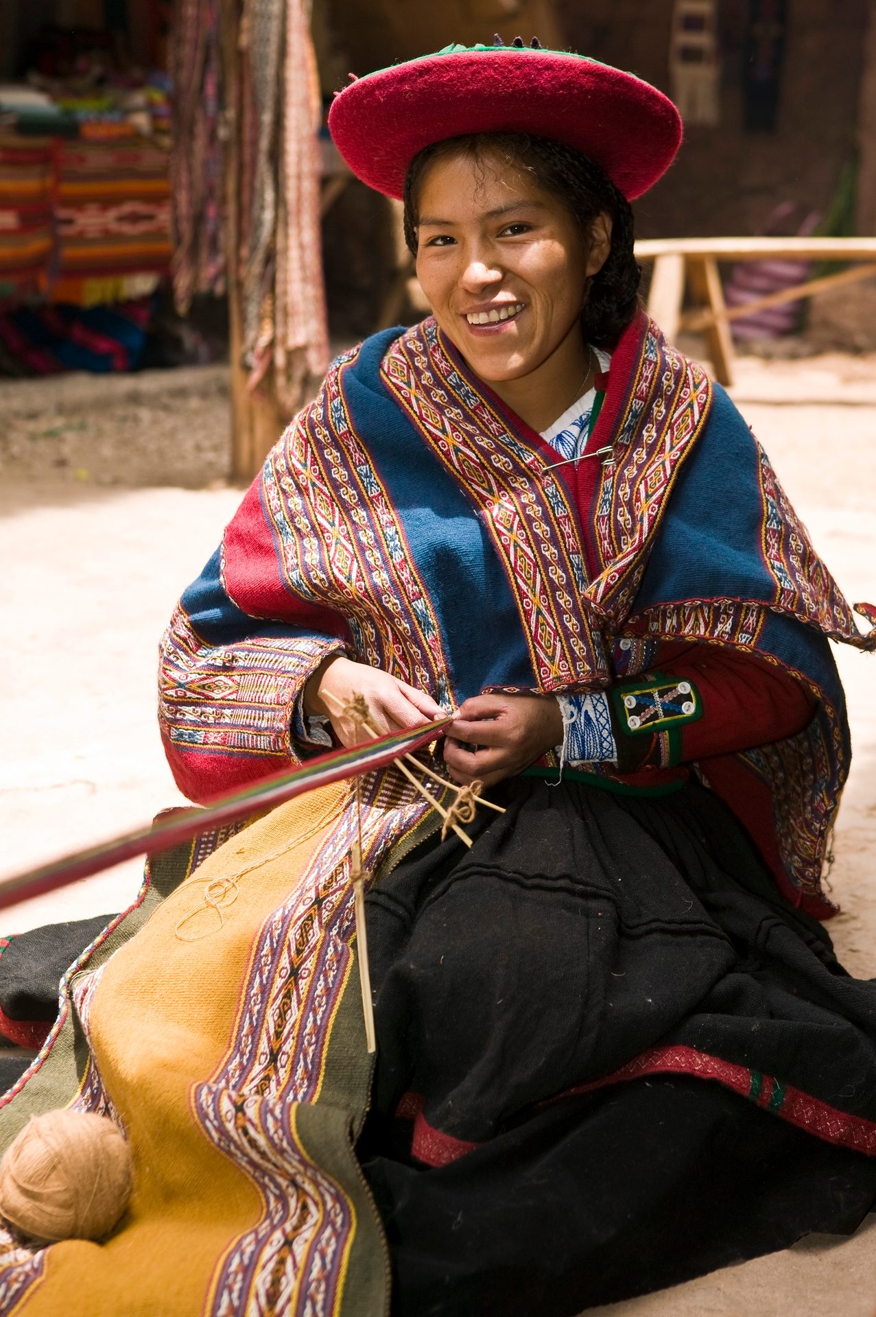 A smiling woman in traditional attire and red hat, handweaving outdoors.