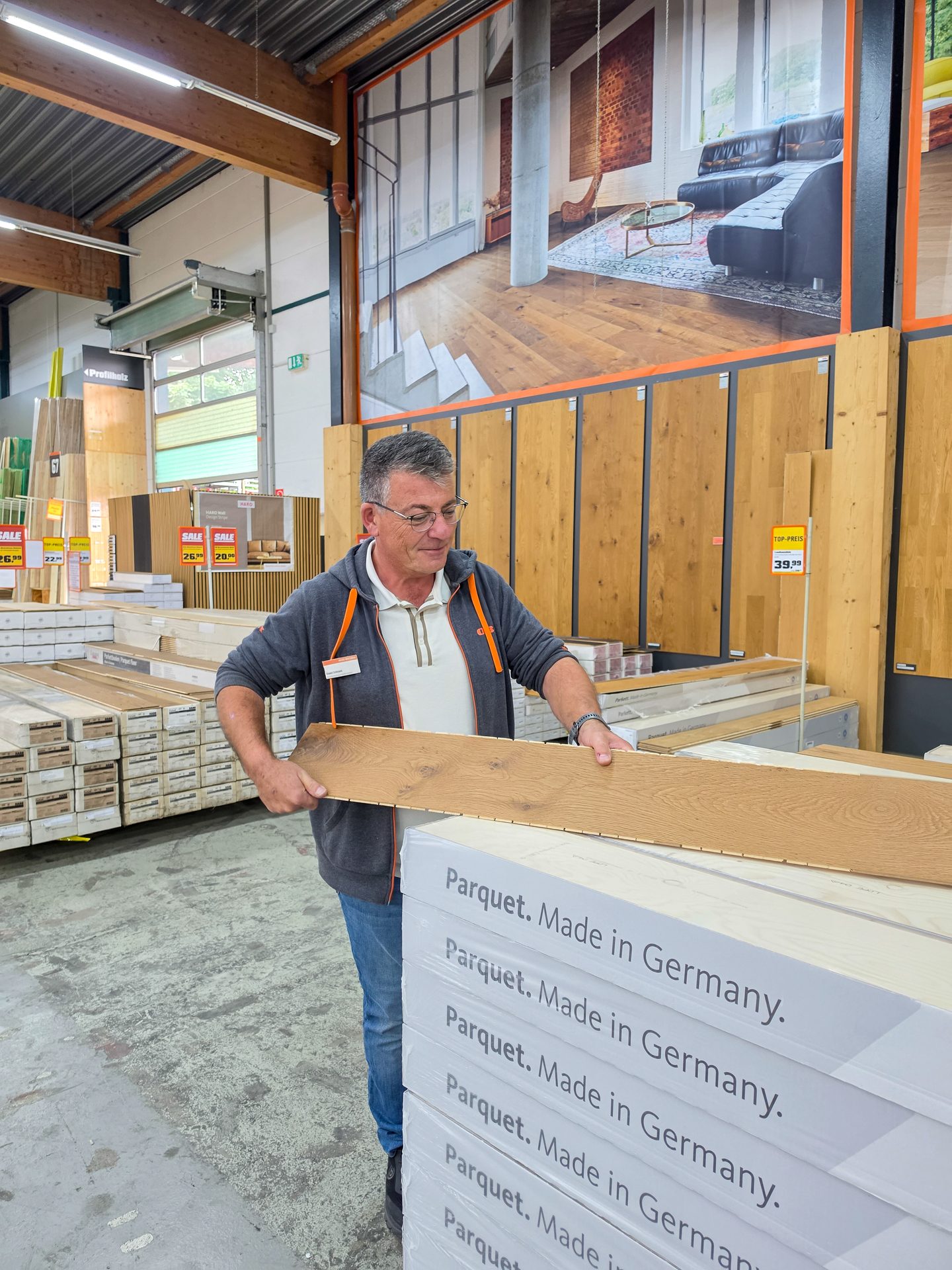 A man in a hardware store examining a "Made in Germany" parquet flooring sample, with stacks of boxes nearby.