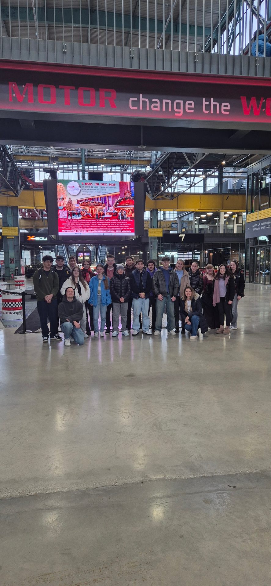 Group of young people indoors, with a large screen behind them and a red sign: MOTOR change the W.