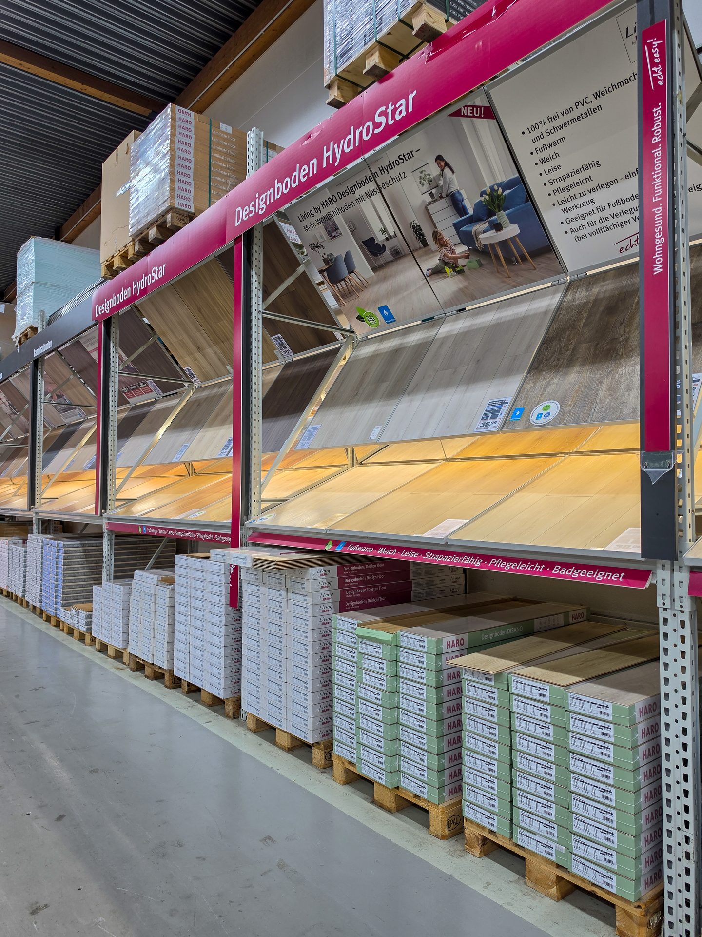 Warehouse display for 'Designboden HydroStar' flooring, showing product features and stacked boxes on pallets.