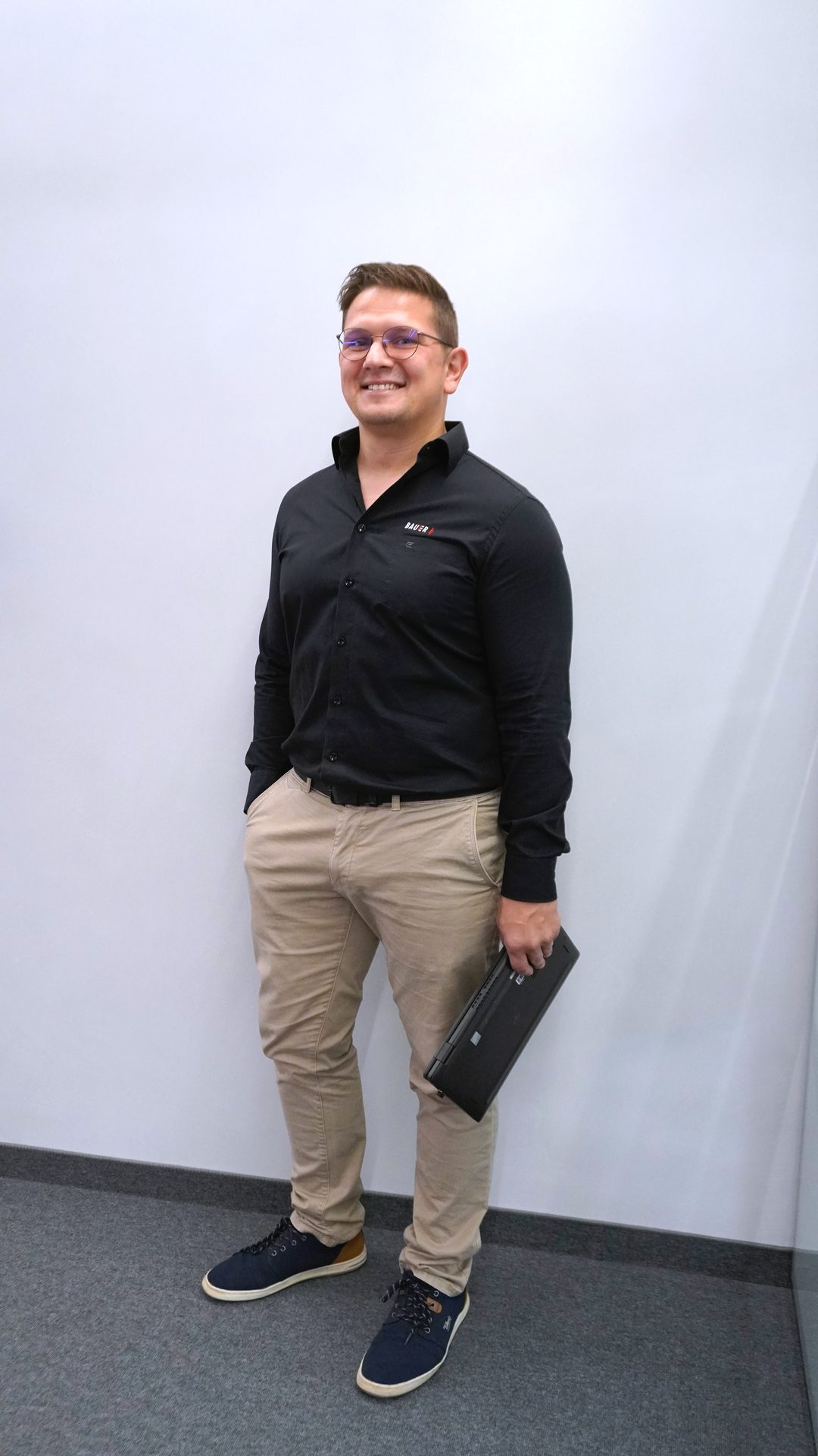 Smiling man in glasses, black shirt, khaki pants, and sneakers, holding a laptop.