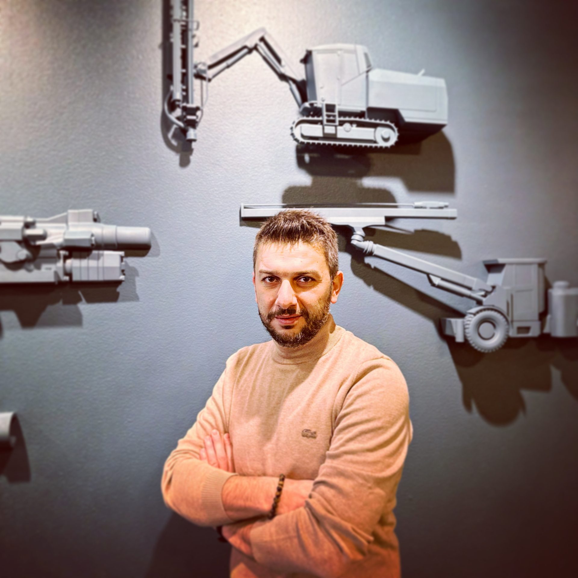 Man in a turtleneck sweater posing against a dark wall with 3D models of construction vehicles.