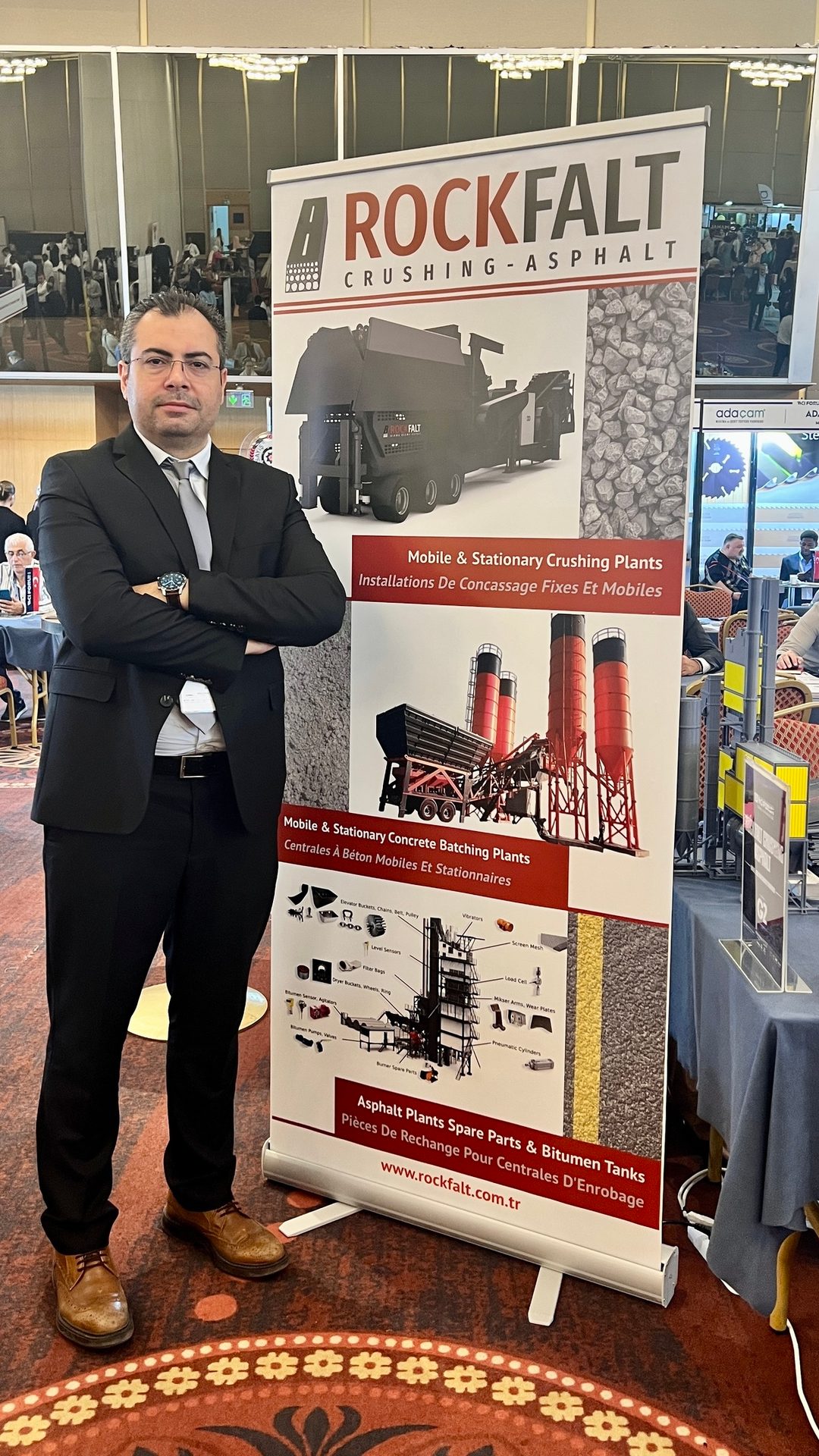 Man in suit standing by a "ROCKFALT" banner advertising crushing and asphalt plants.