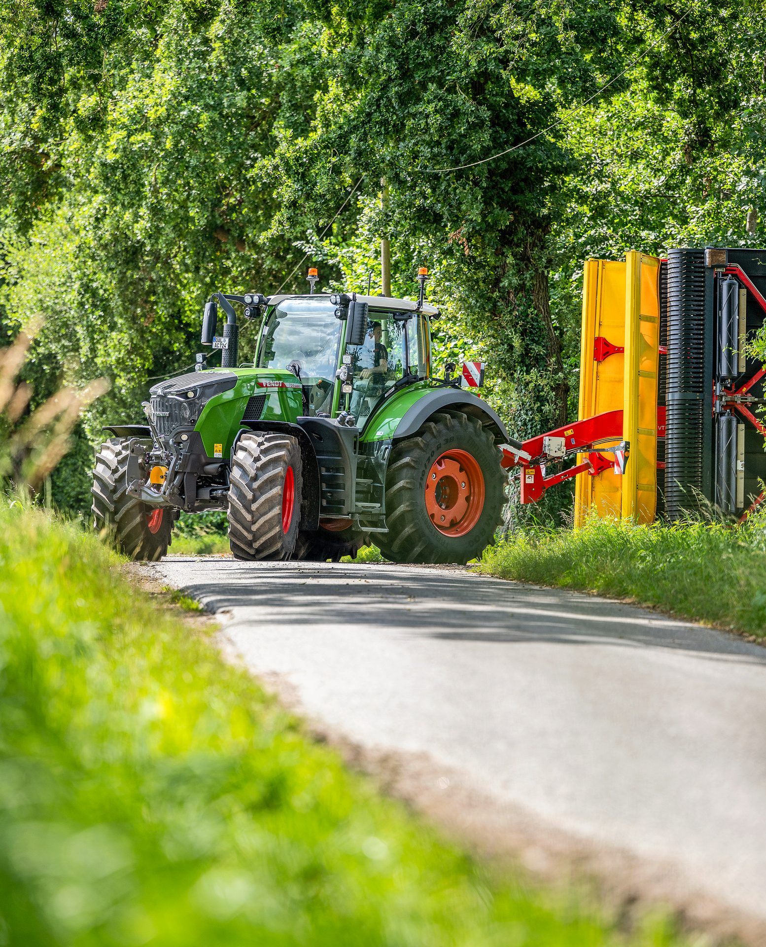 A green Fendt tractor with a yellow agricultural implement drives on a road surrounded by lush greenery.