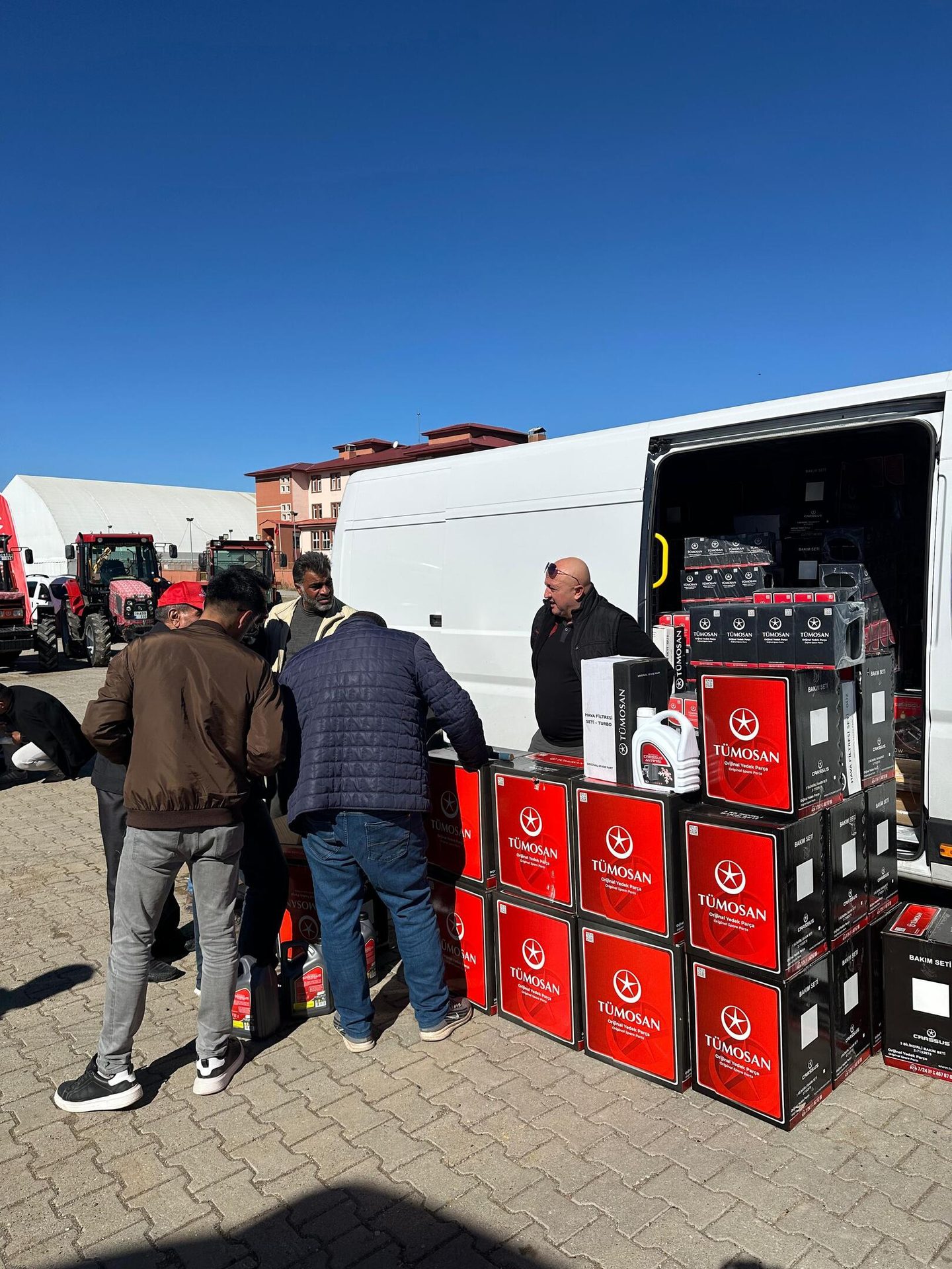 Men distribute TÜMOSAN tractor parts and oil from a van, with tractors and buildings in the background.
