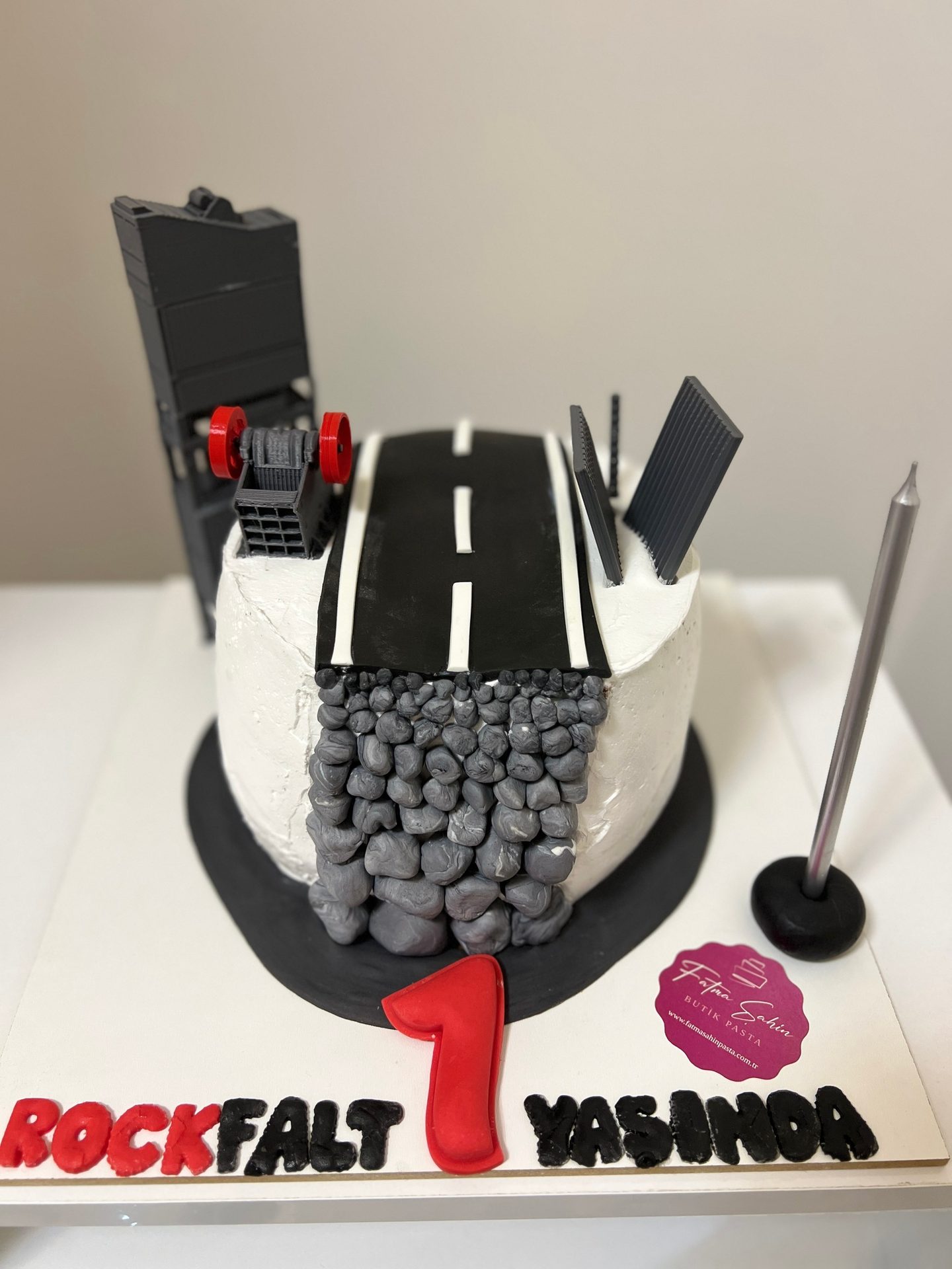 A construction-themed birthday cake featuring a road, rock-like decorations, a "1", and "ROCKFALT YASIMDA" text.