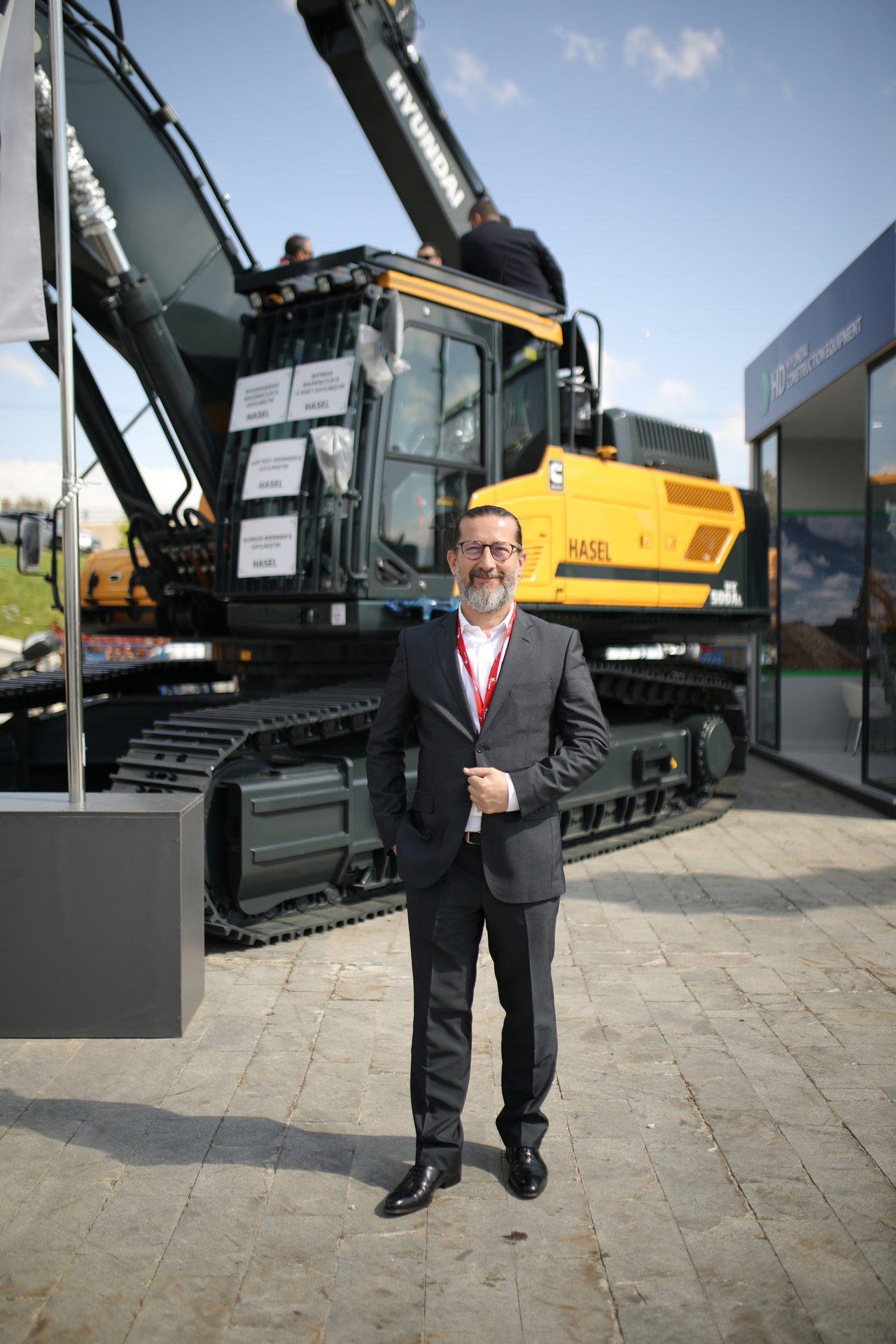 Man in a business suit and glasses posing in front of a large yellow Hyundai excavator.
