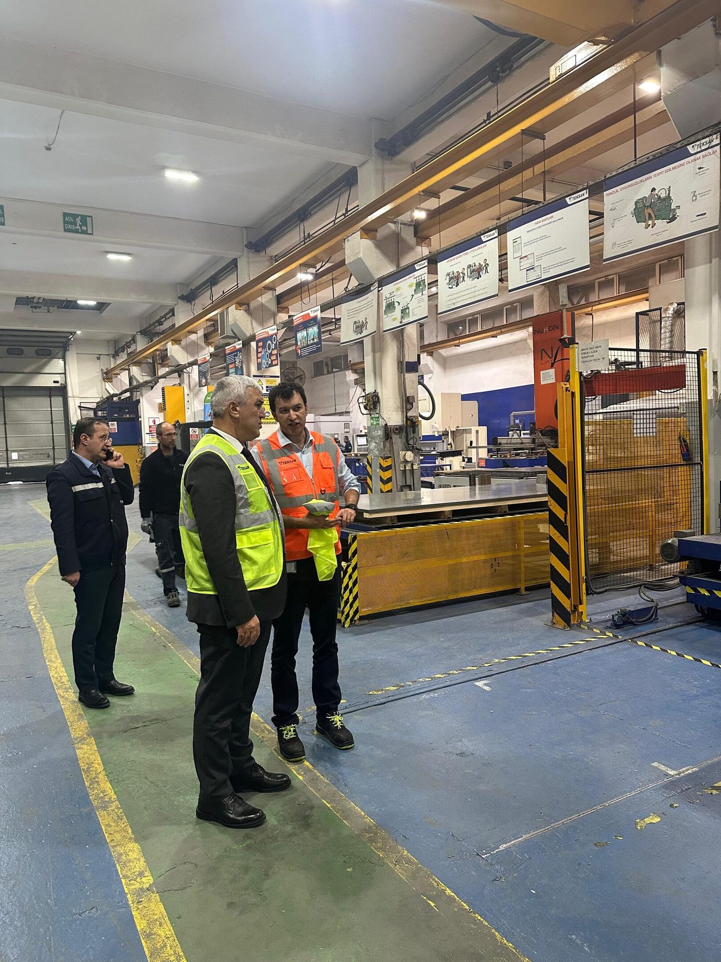 Three men, two in high-vis vests, talking in a factory with machinery and overhead cranes.