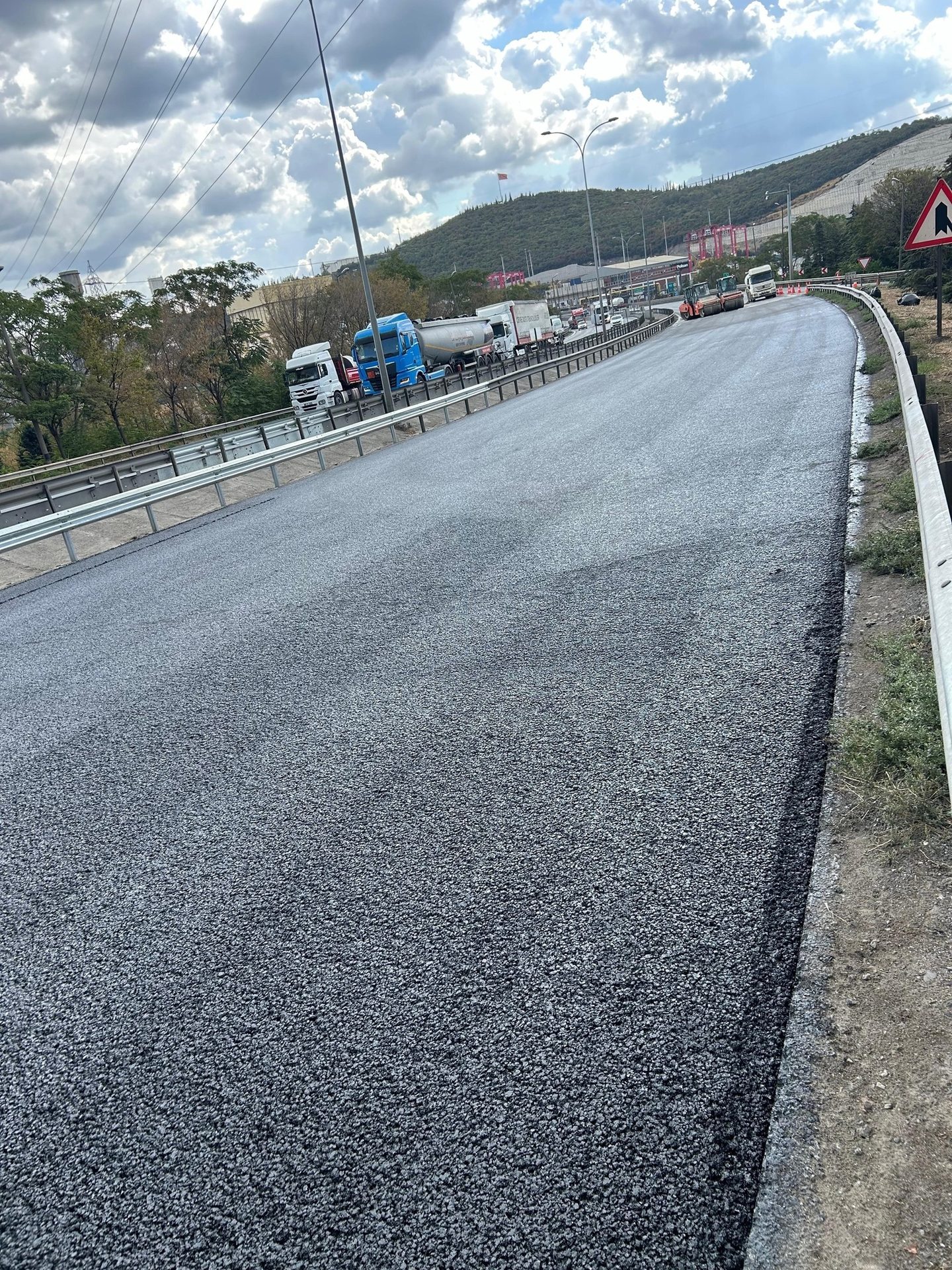 Newly paved asphalt road on an elevated highway, with trucks and hills in the background.
