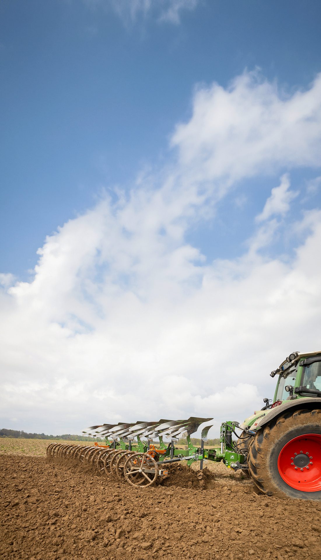 A tractor with a large plow tilling a brown field under a cloudy blue sky.