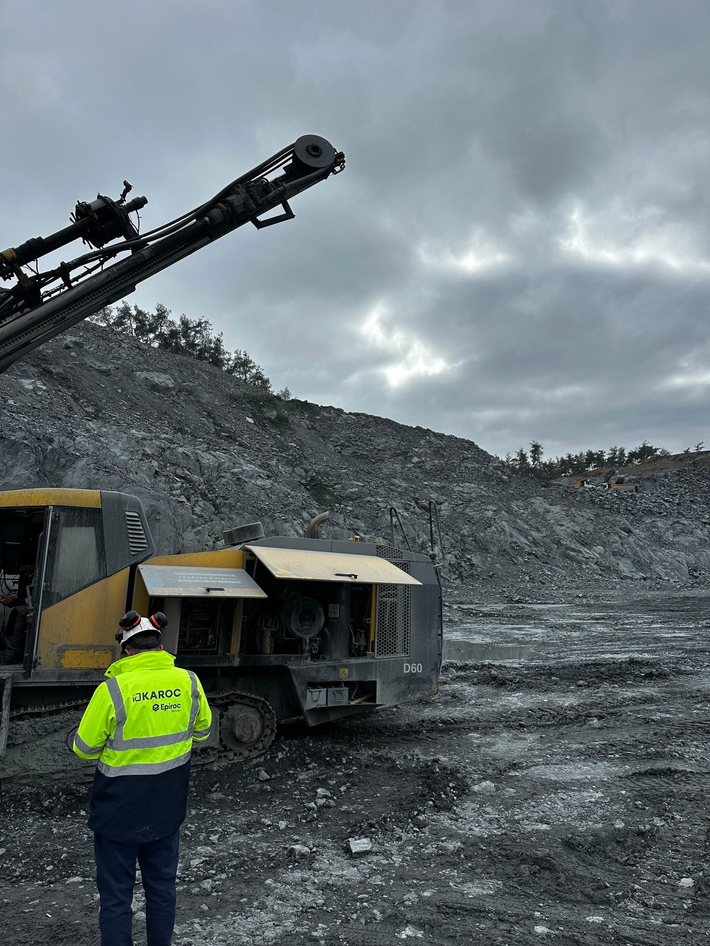 Worker in high-vis jacket stands before heavy drilling rig in rocky quarry under cloudy sky.