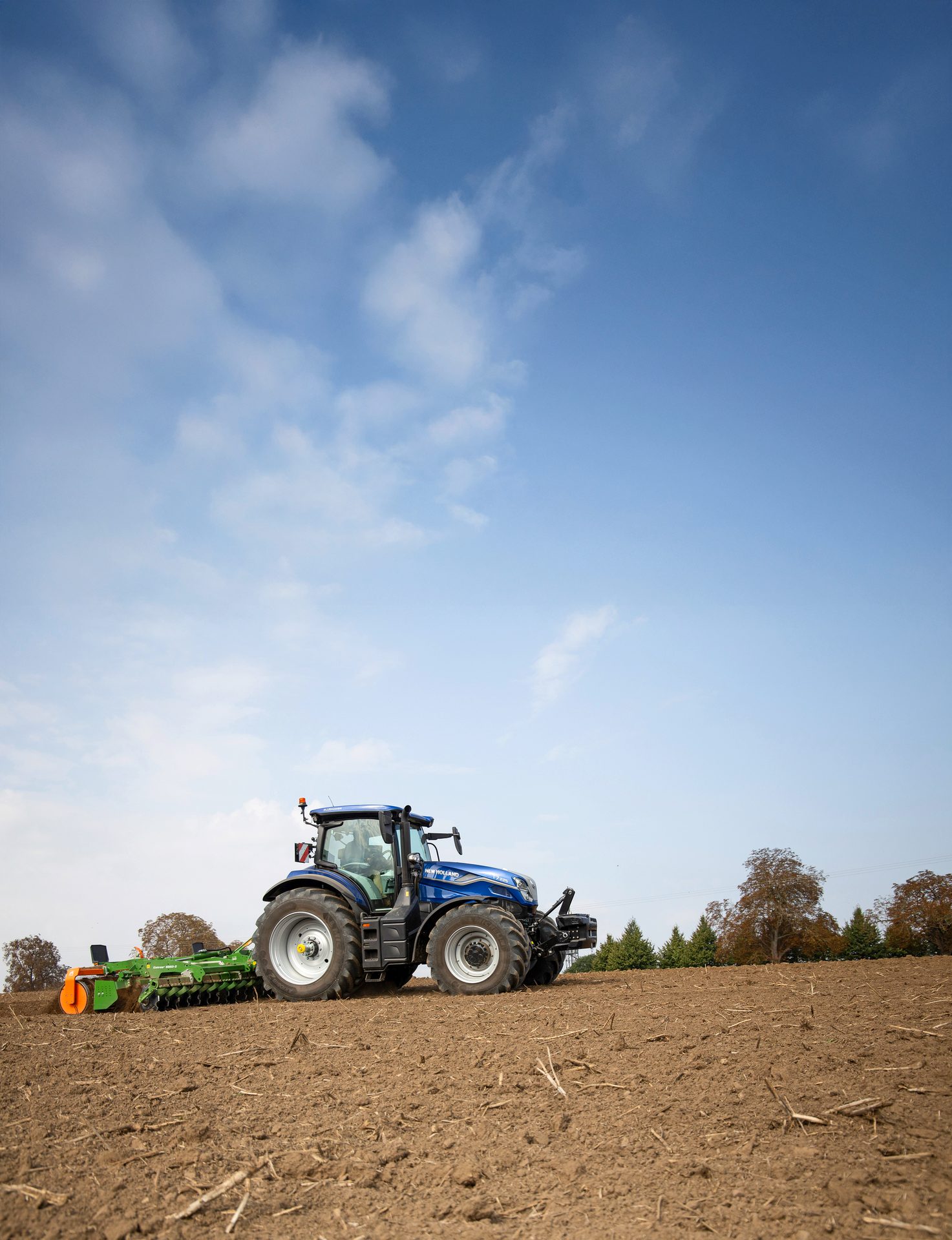 Blue New Holland tractor with a green implement in a field under a blue sky, preparing soil.