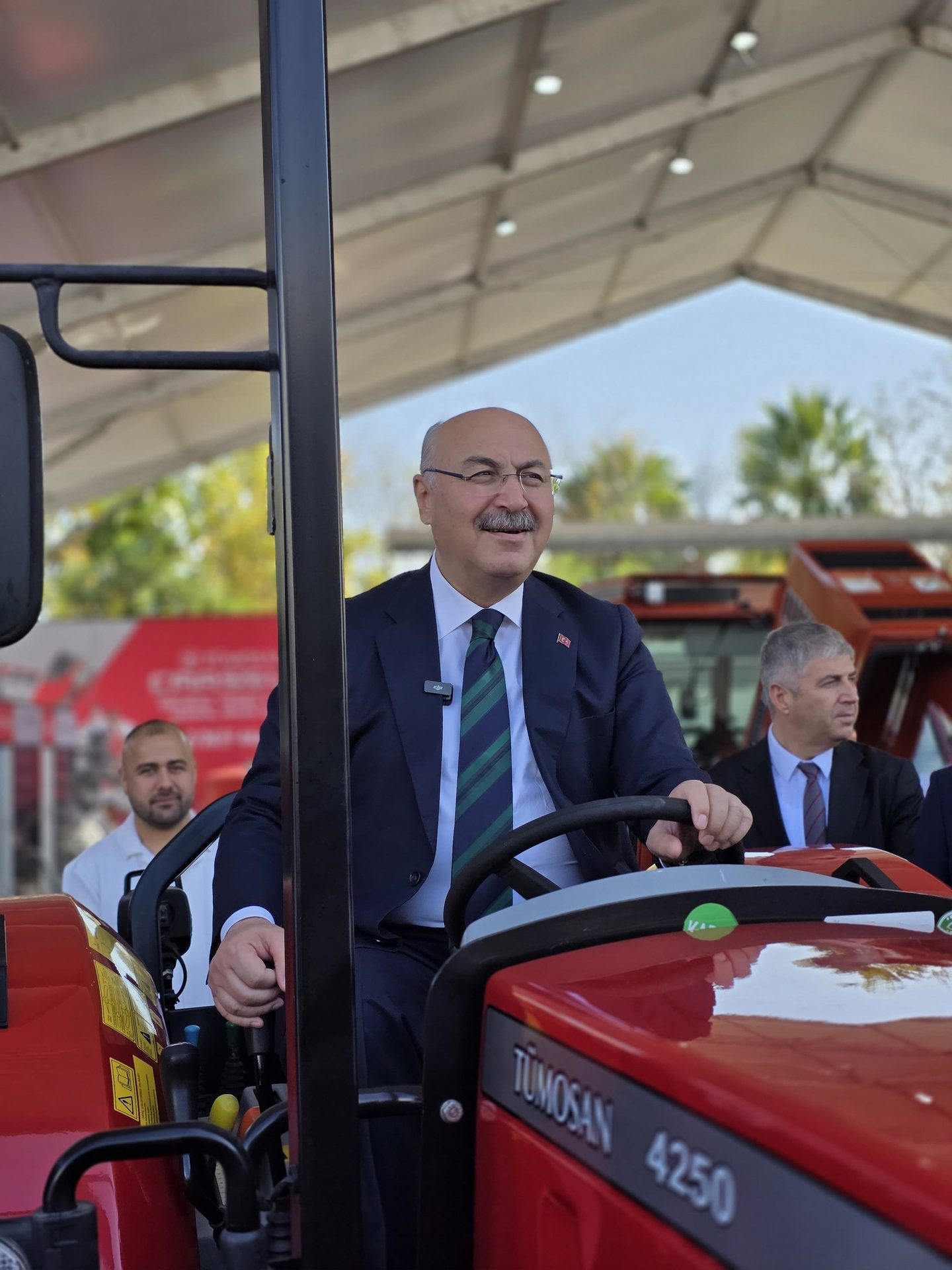 Smiling man in suit drives a red TUMOSAN 4250 tractor, with others nearby in an event setting.