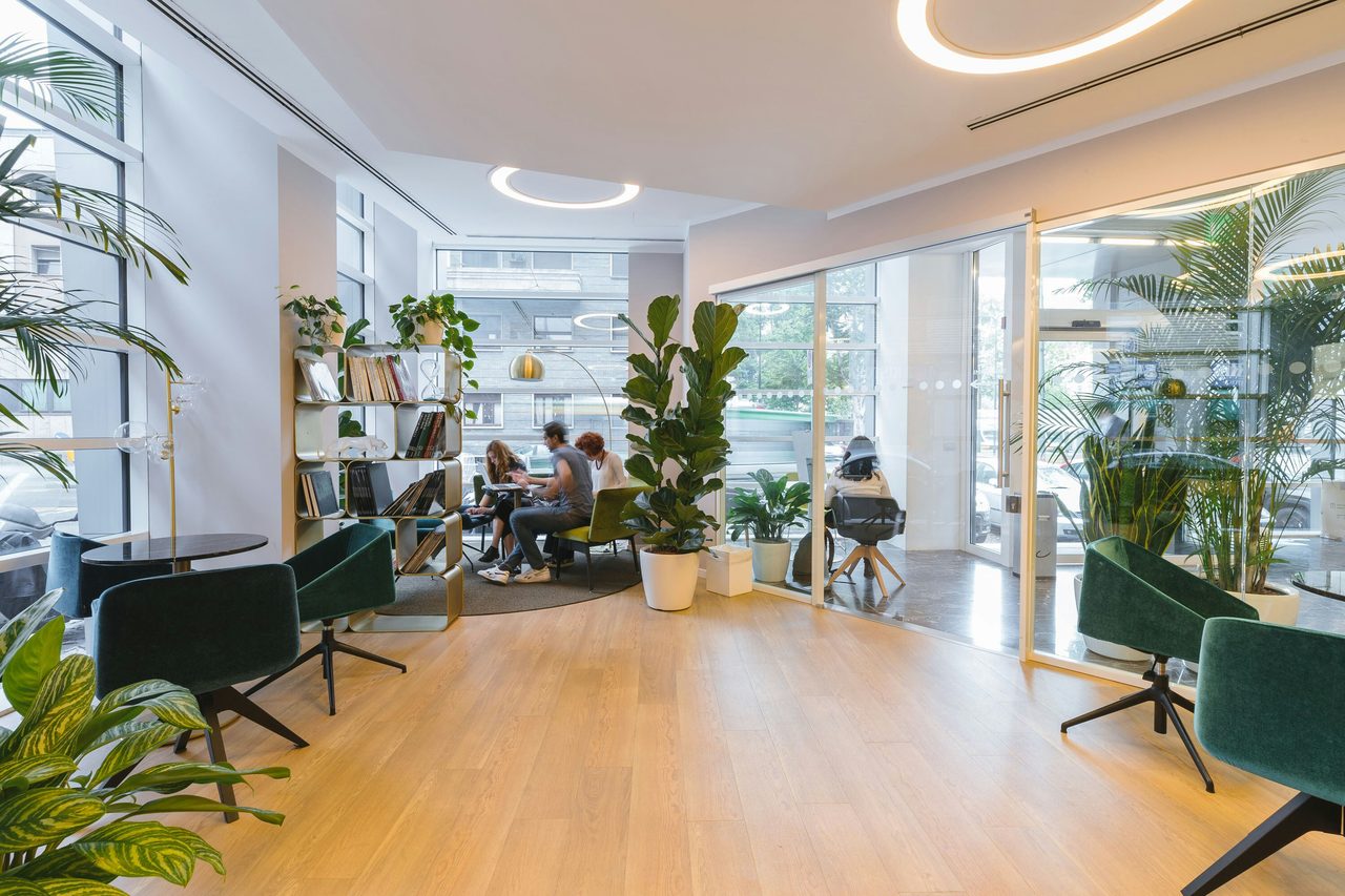 Modern office lounge with people working at a table, large plants, bookshelves, and green chairs.