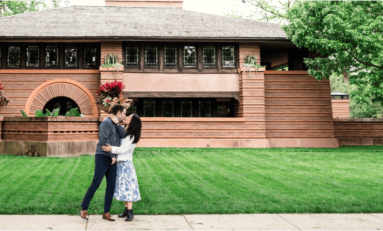 A couple kisses on a sidewalk in front of a brown, Frank Lloyd Wright-style house with green grass.