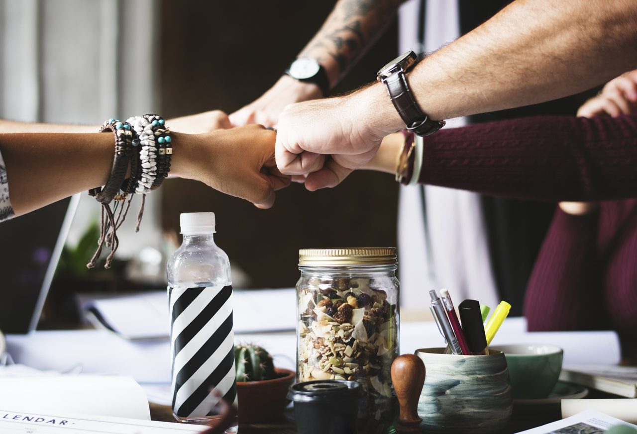 Close-up of four people's fists bumping together over a desk with office supplies, symbolizing teamwork.