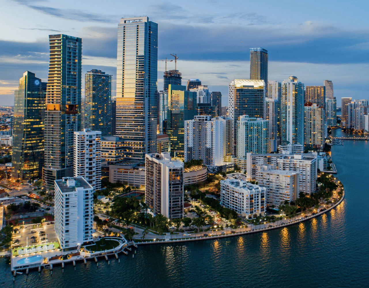 Aerial view of an illuminated city skyline with skyscrapers at dusk, along a waterfront.