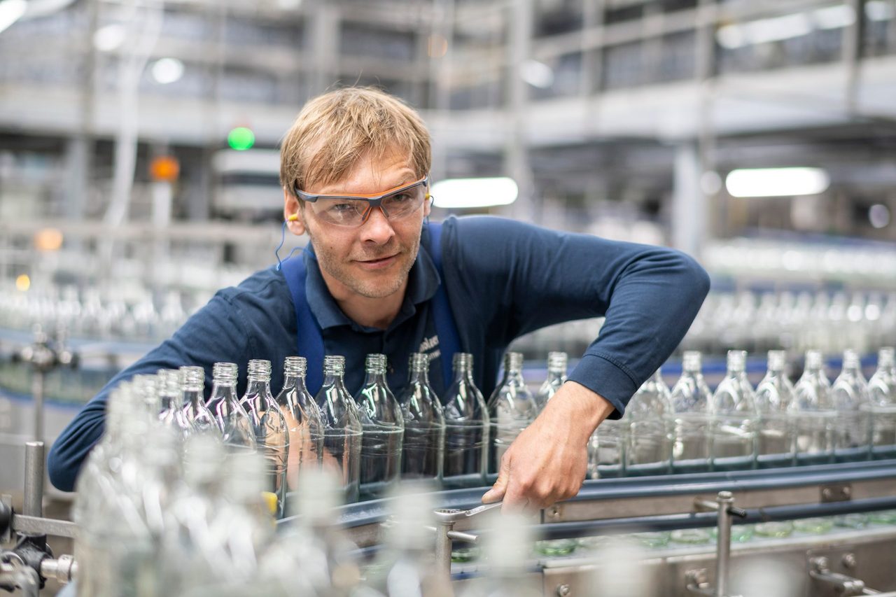 Factory worker with safety glasses adjusting bottles on a production line.