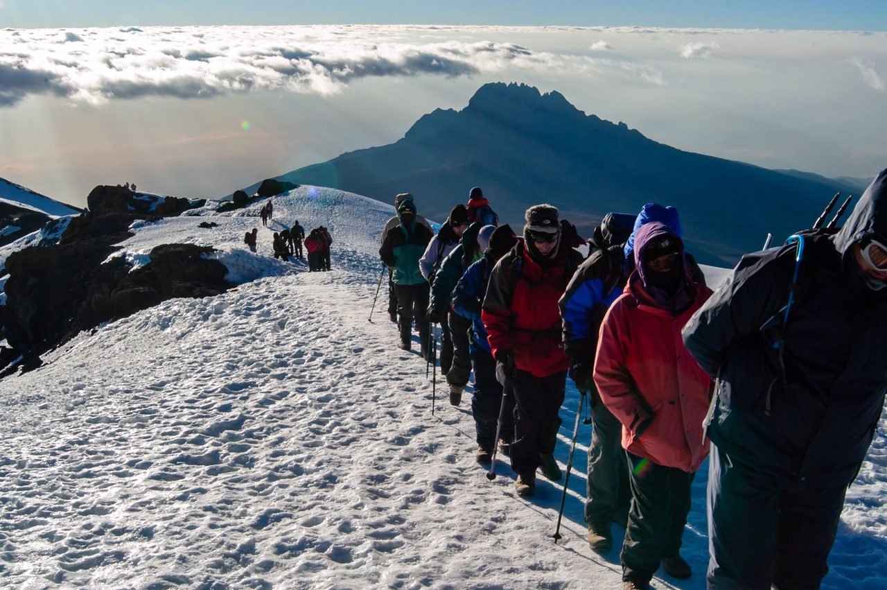 Final ascent to the top of Kilimanjaro or Uhuru Peak. A line of hikers stepping over snow. Mount Mawenzie in the distance looking down on the clouds.