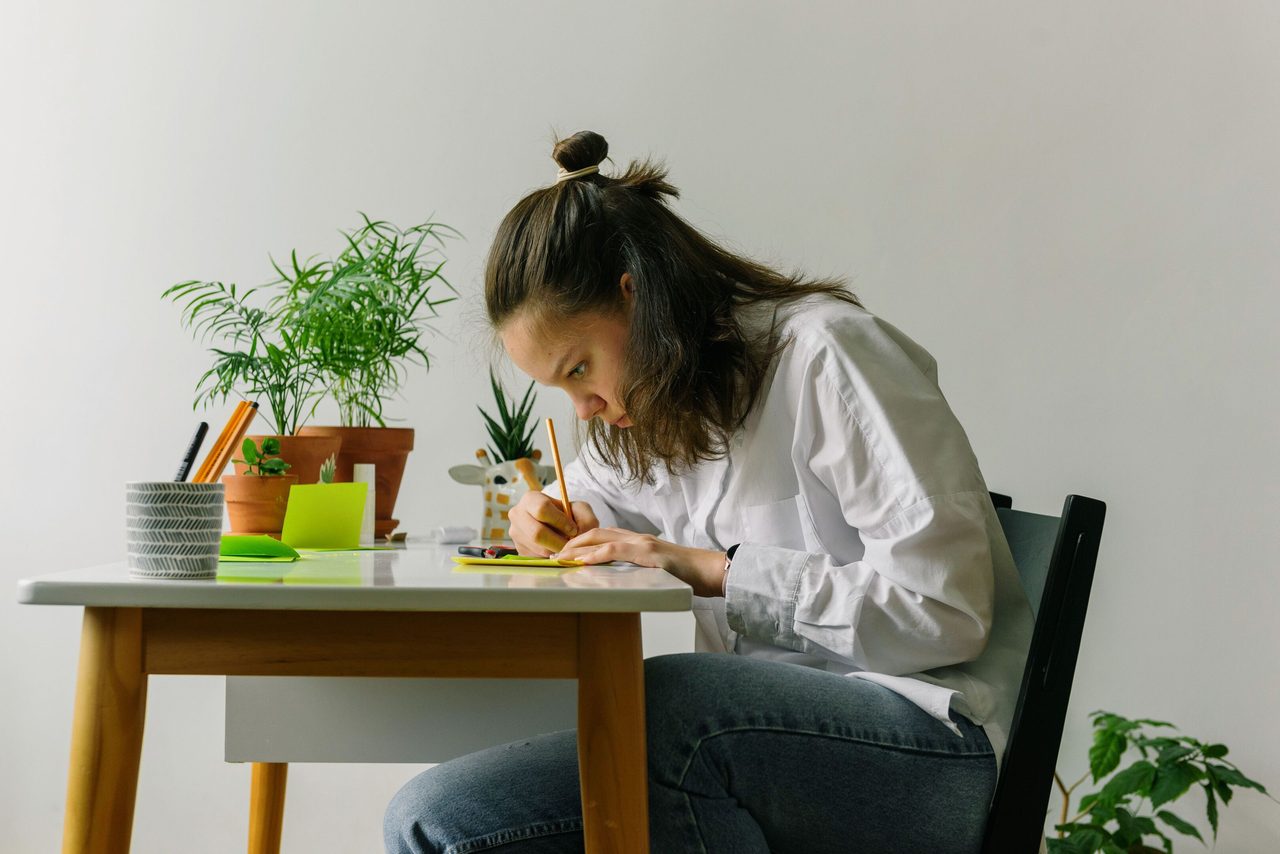 A young woman intensely writing at a desk surrounded by potted plants indoors.. Ksenia Chernaya: https://www.pexels.com/@kseniachernaya