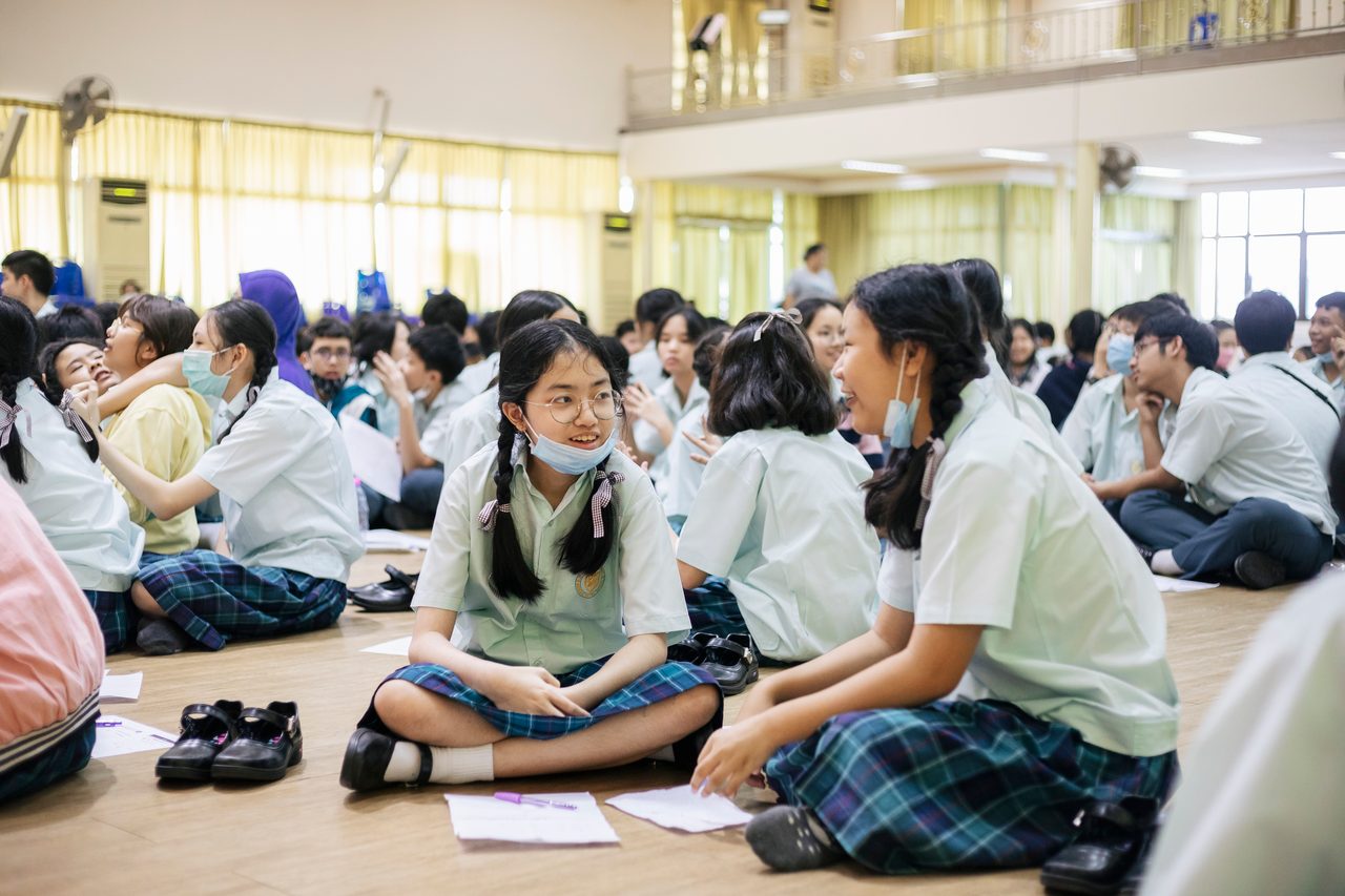 Two young Thai students in school uniform sitting on the floor of a gymnasium, talking to each other