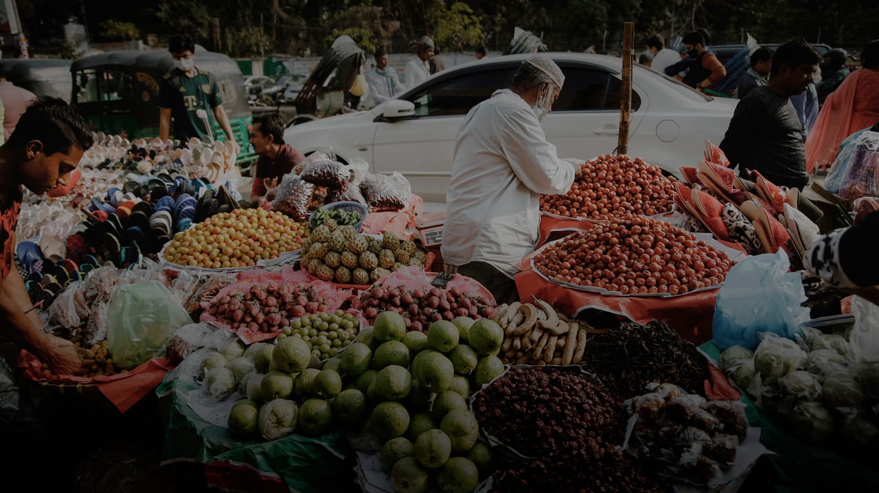 Photo of a market stall with baskets overflowing of fruit, vegetables and berries.  In the middle of the basket stands an elderly vendor in a white shirt. 