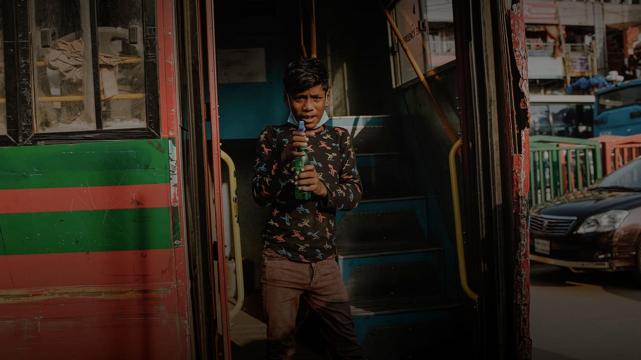 Photo of a young bangladeshi boy standing on bus-steps, pointing a spray bottle at the camera