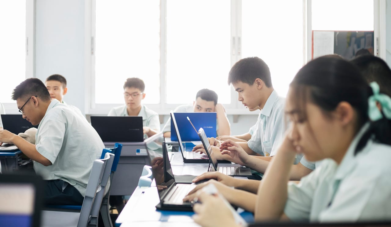 Photo of young uniformed thai pupils sitting on their desks in a brightly lit classroom, intently working on their computers.