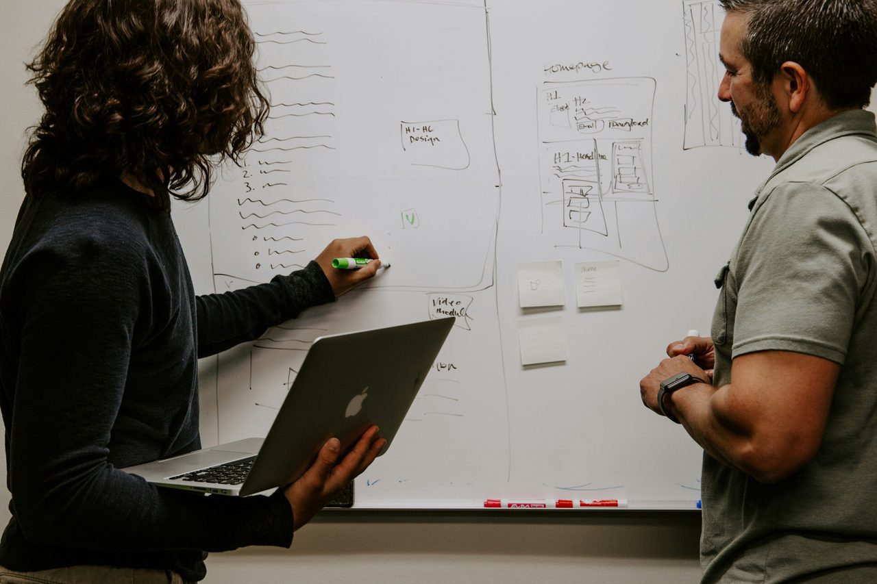 Two people collaborating on a whiteboard, one writing diagrams and notes while holding a laptop, the other observing.