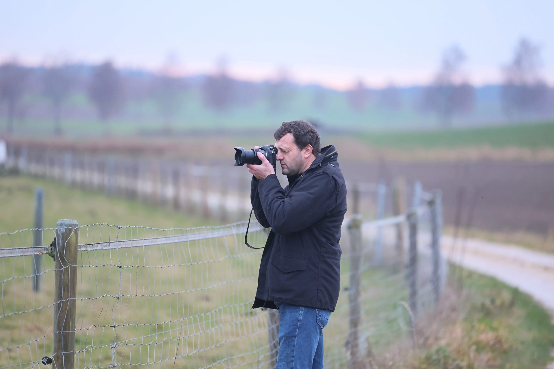 A man in a black jacket photographs outdoors with a camera, next to a fence.