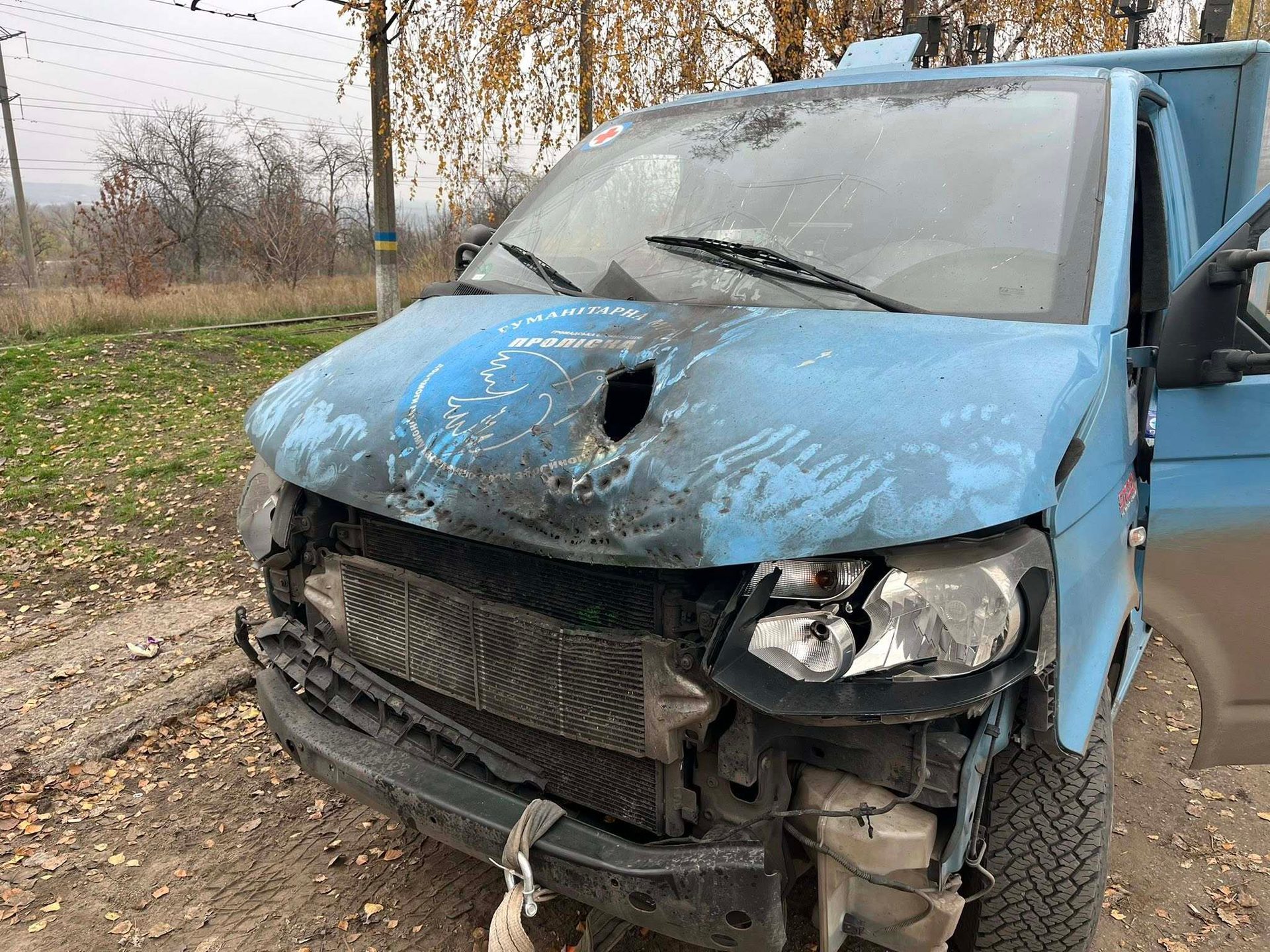Severely damaged blue humanitarian aid van with a large hole in its hood and a broken front.