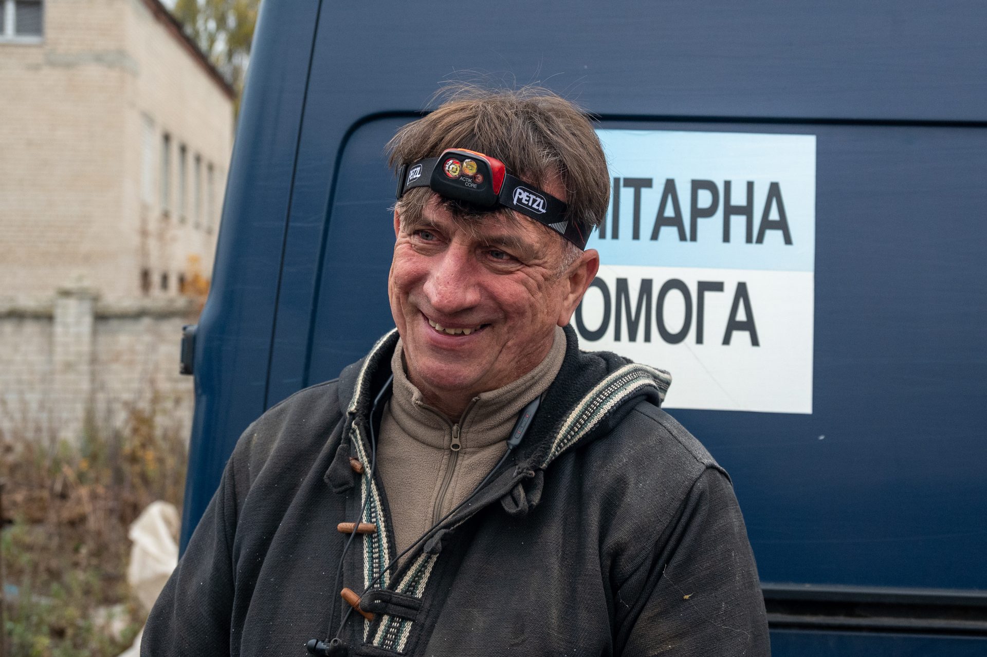 A smiling man wearing a Petzl headlamp and a jacket, with a blue humanitarian aid van in the background.
