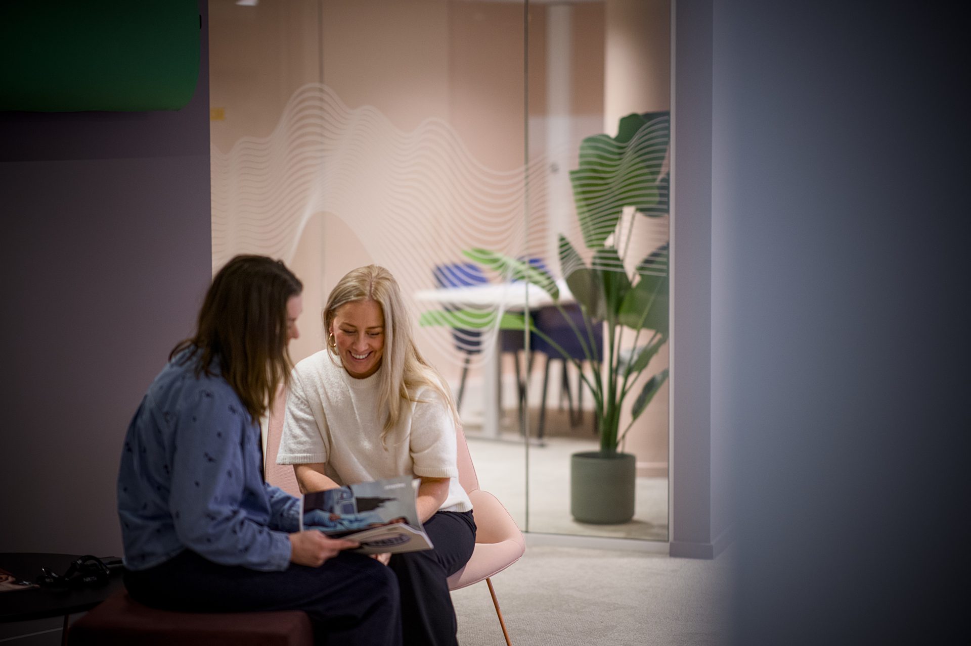 Two women smiling, looking at a magazine in a modern office with a glass wall.