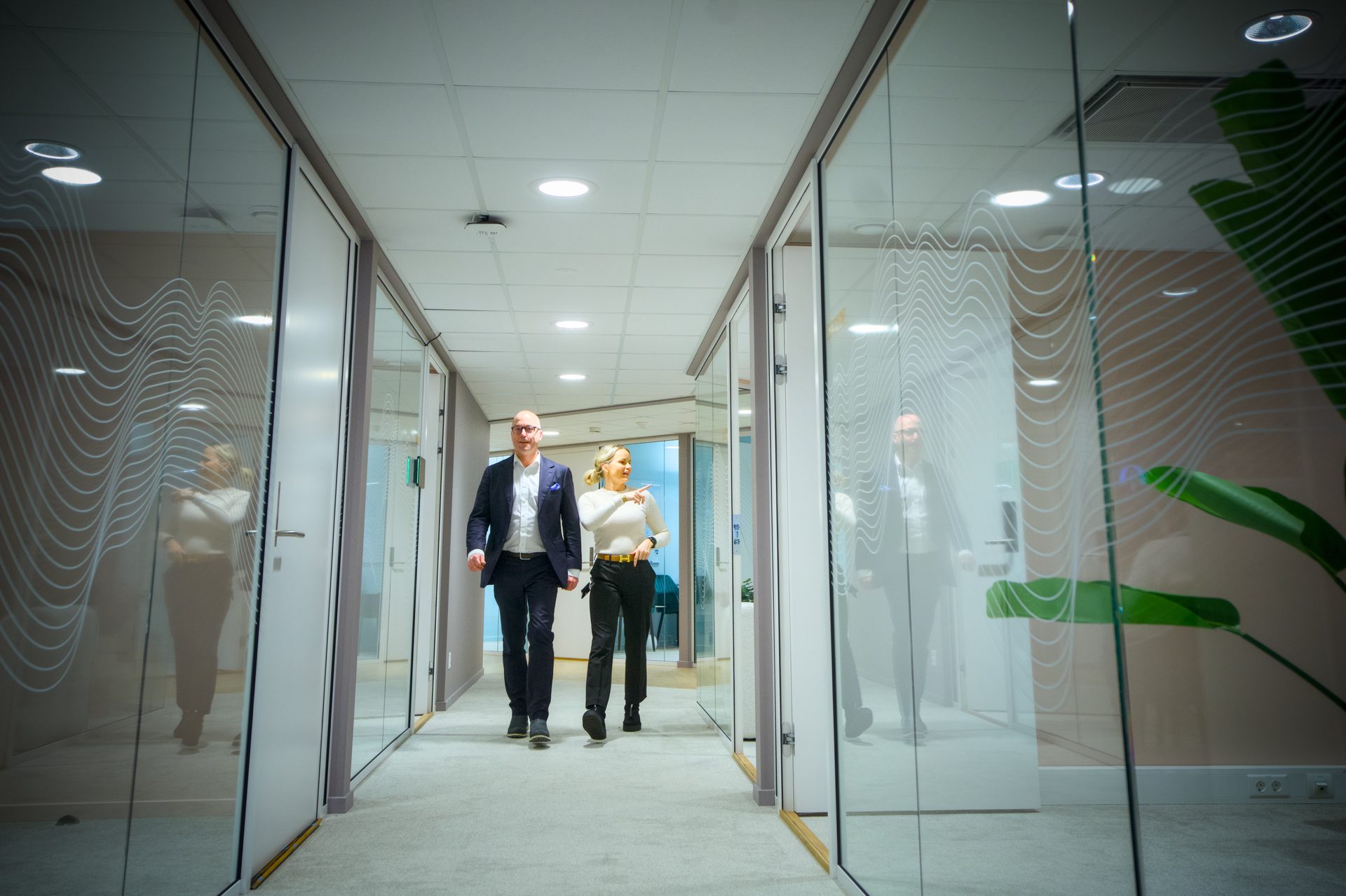 Man and woman walk in a bright office hallway with glass walls, woman pointing.
