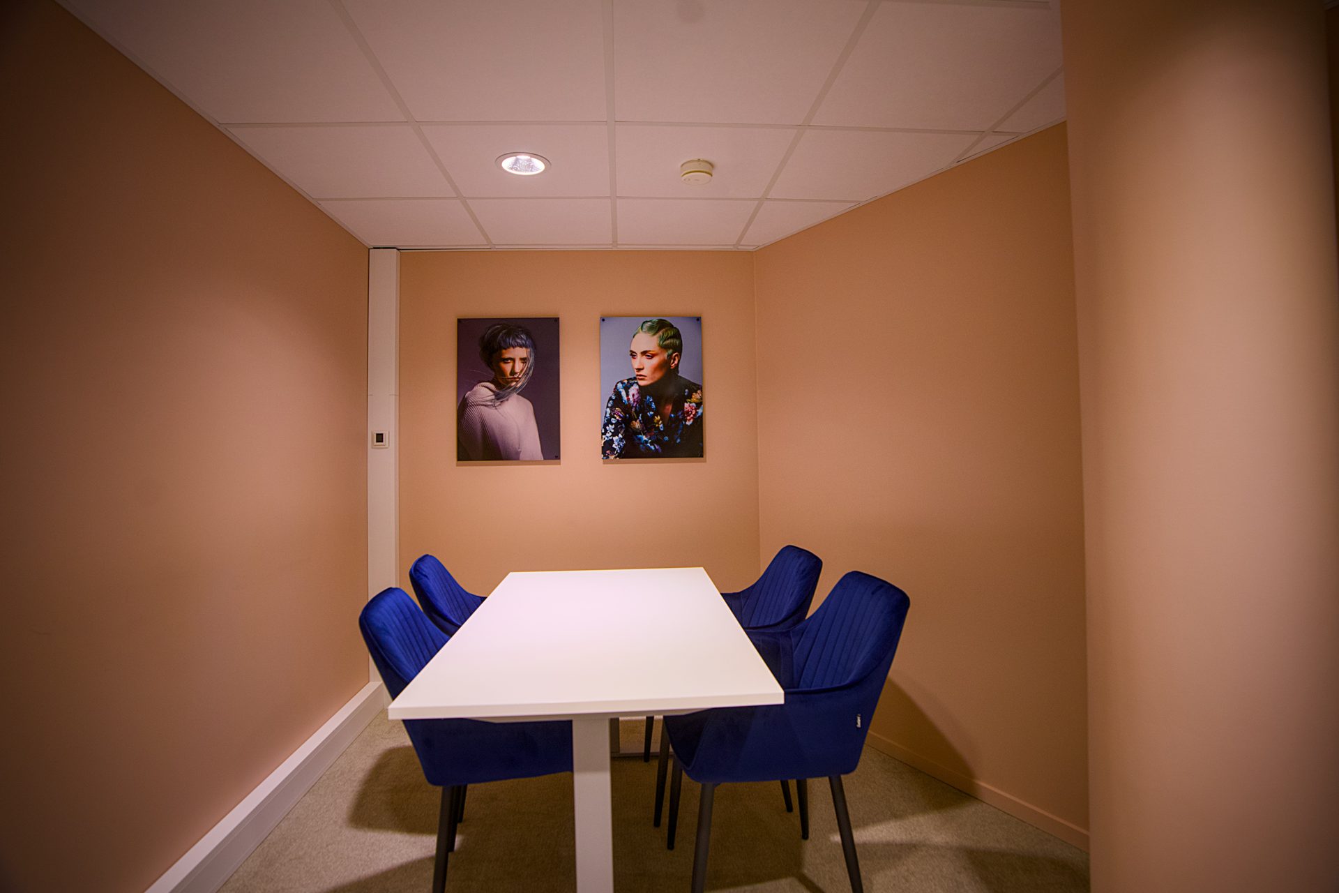 A small room with peach walls, a white table, four blue chairs, and two wall portraits.