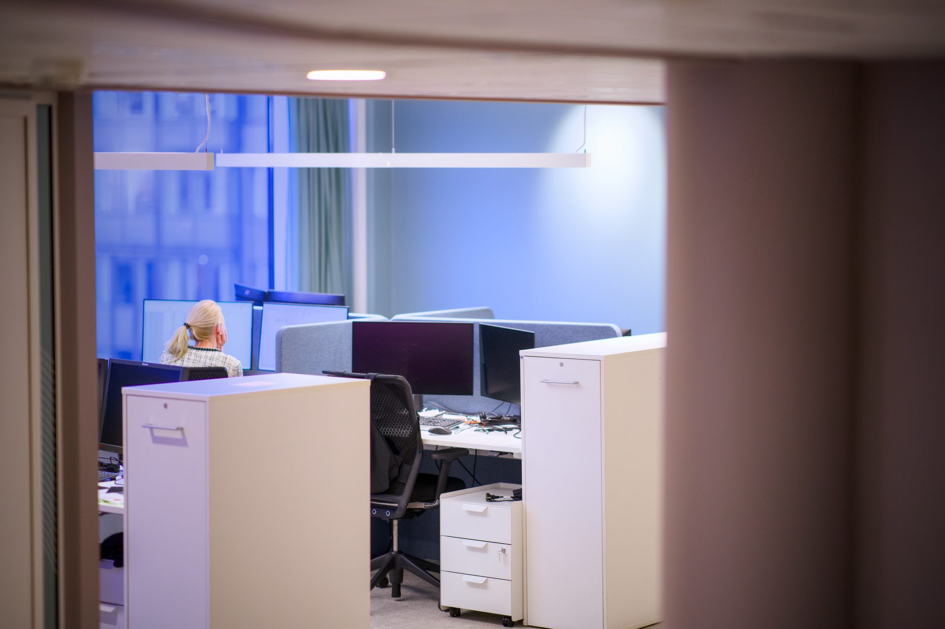 Person working at a computer in a blue-lit office, seen through a doorway.