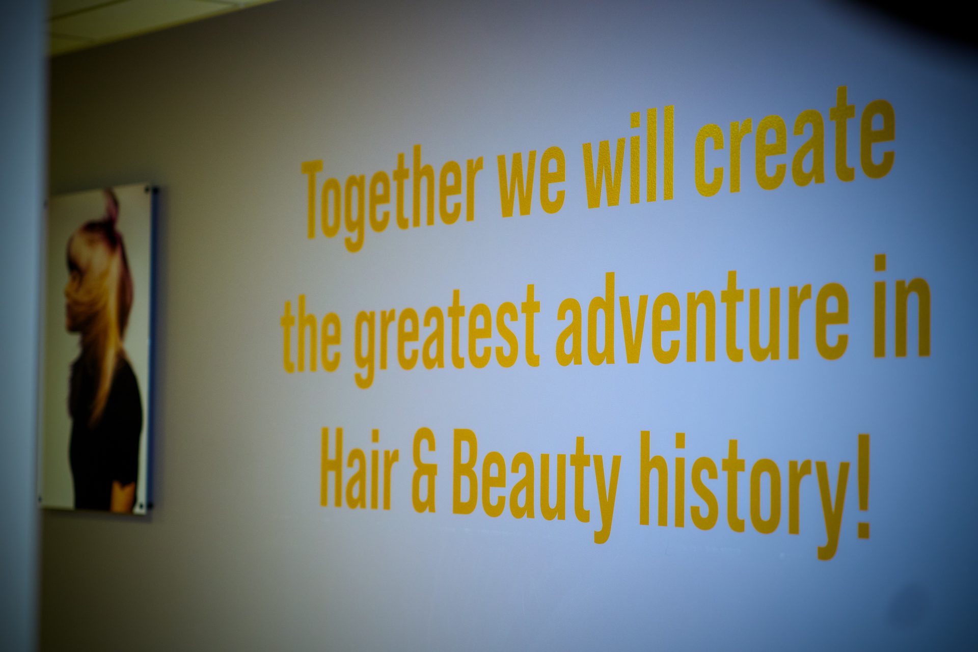 Yellow text on a wall: "Together we will create the greatest adventure in Hair & Beauty history!". Blurry image.