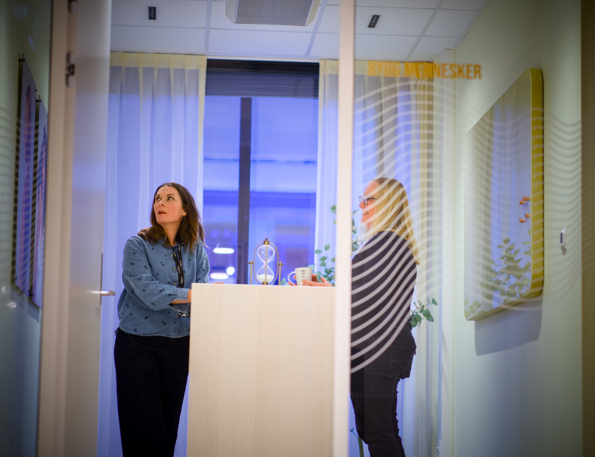 Two women interact in a bright office, separated by a glass partition, with an hourglass on a counter.