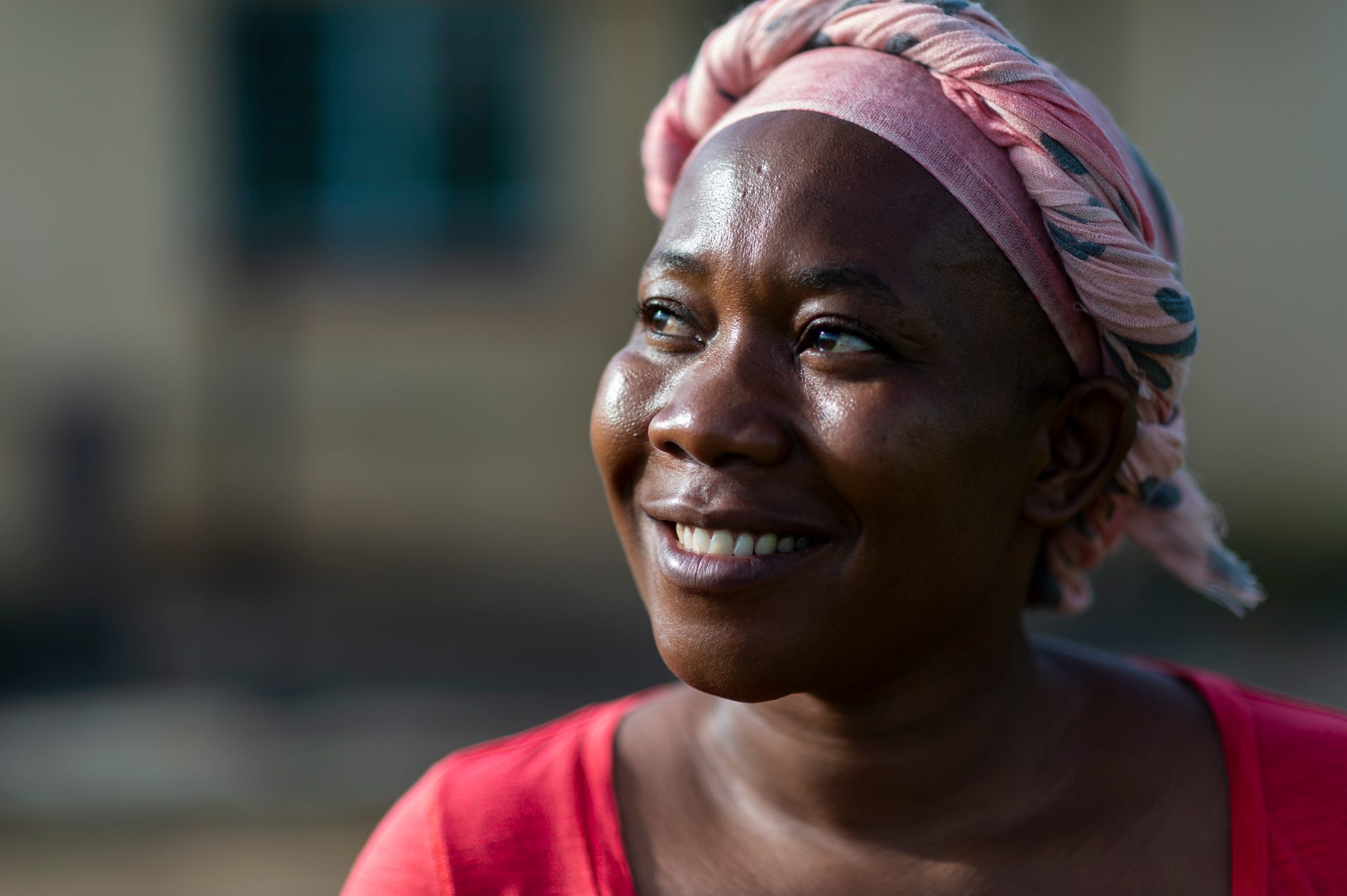Image depicts an african woman smiling and looking off in to the distance. She is a breast cancer survivor wearing a pink hair wrap.