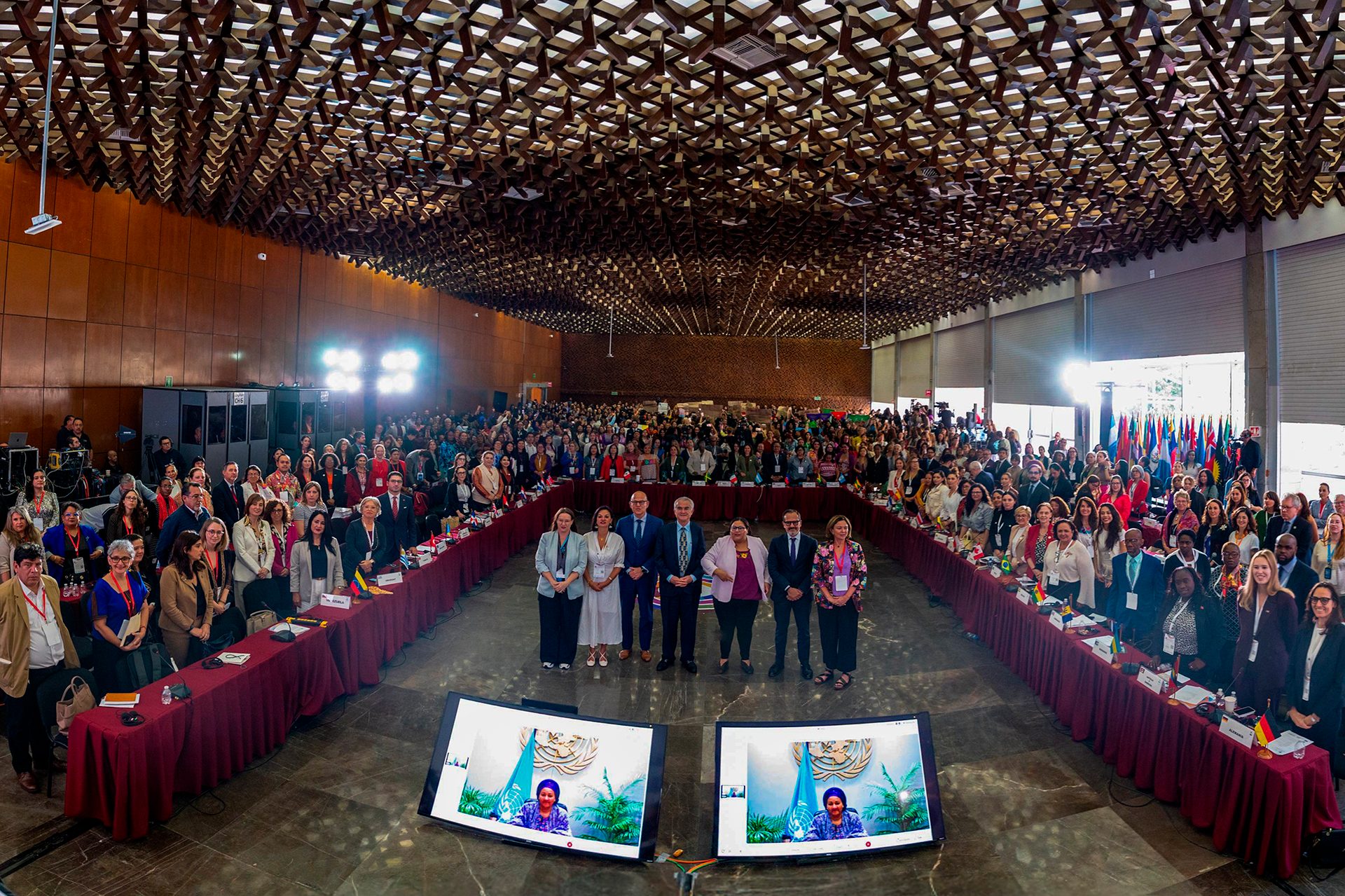 Wide shot: large conference with diverse attendees, a panel, and screens showing a virtual speaker.